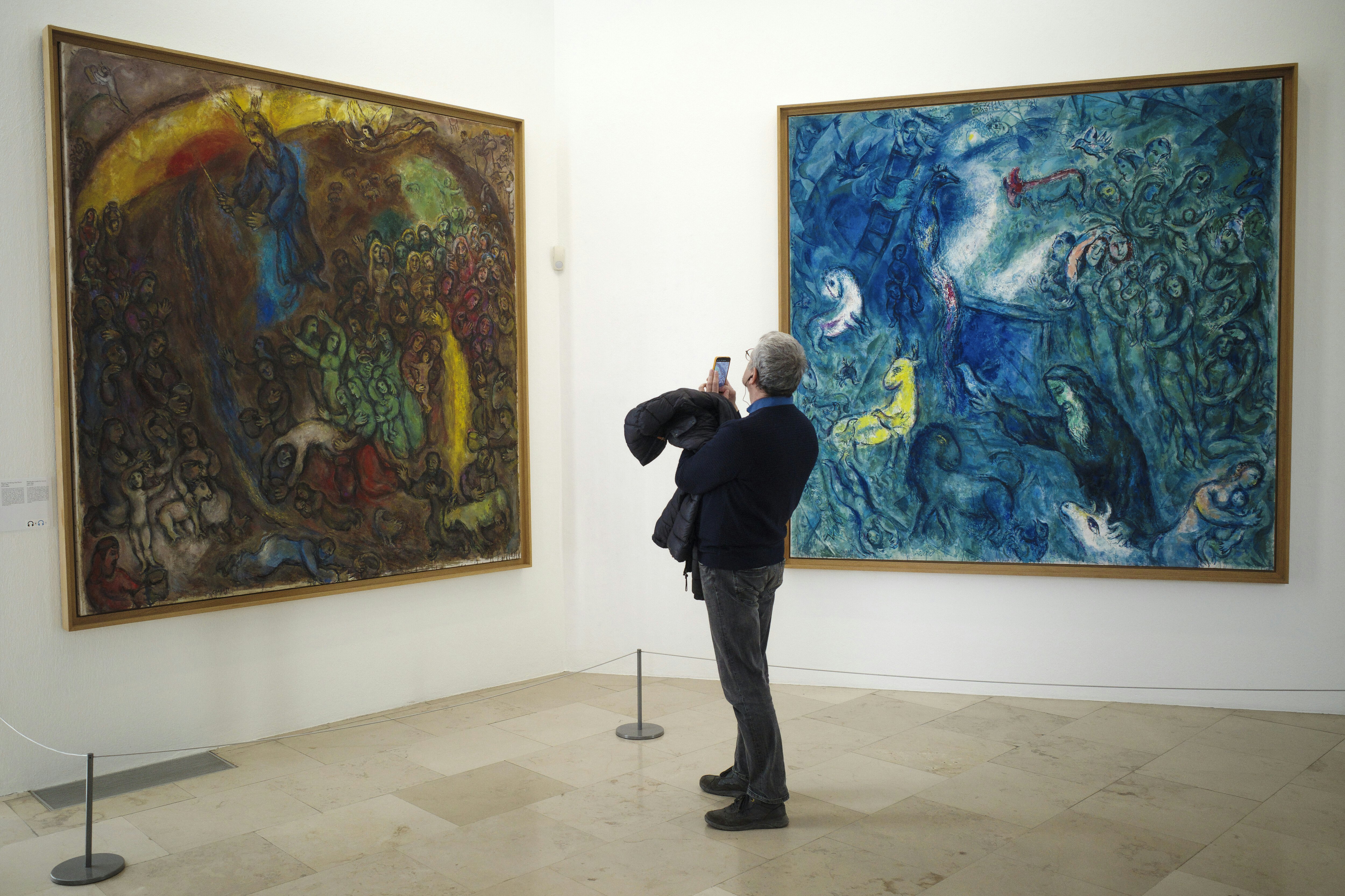 Vyras stovi galerijoje ir naudoja savo telefoną, kad nufotografuotų meno kūrinį. Du dideli paveikslai - vienas raudonas, auksinis ir kitas spalvas; Kitas dažniausiai mėlynais tonais - pakabinkite ant baltų sienų.