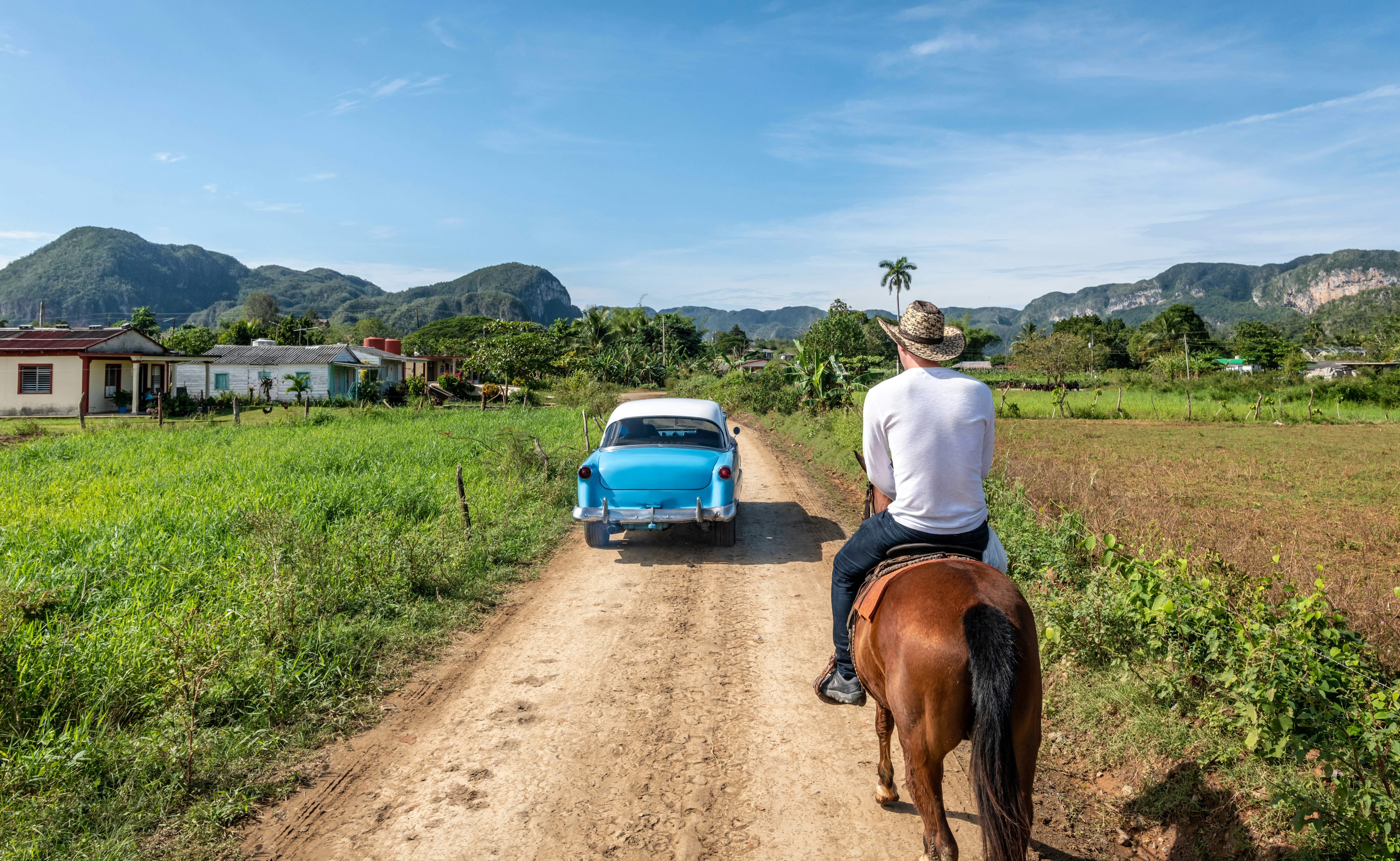 Mėlynas klasikinis automobilis praeina kaubojų nešiojamą vyrą ant arklio ant purvo kelio, vedančio į Vinales, Kubą
