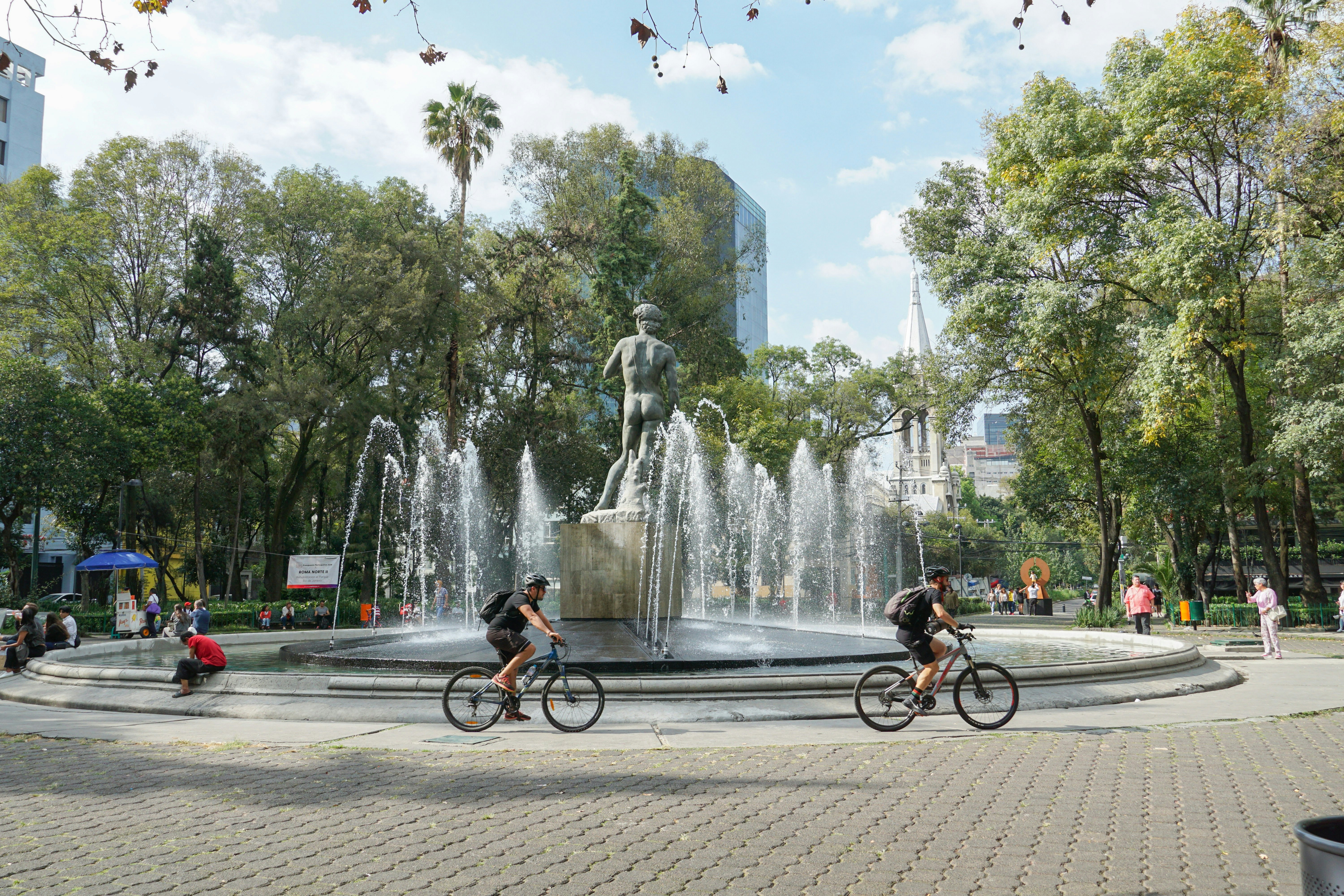 Jaunimas važiuoja dviračiais aplink didelį apskritą fontaną su Mikelandželo Davido kopija centre, Plaza Rio de Janeiro, CDMX.