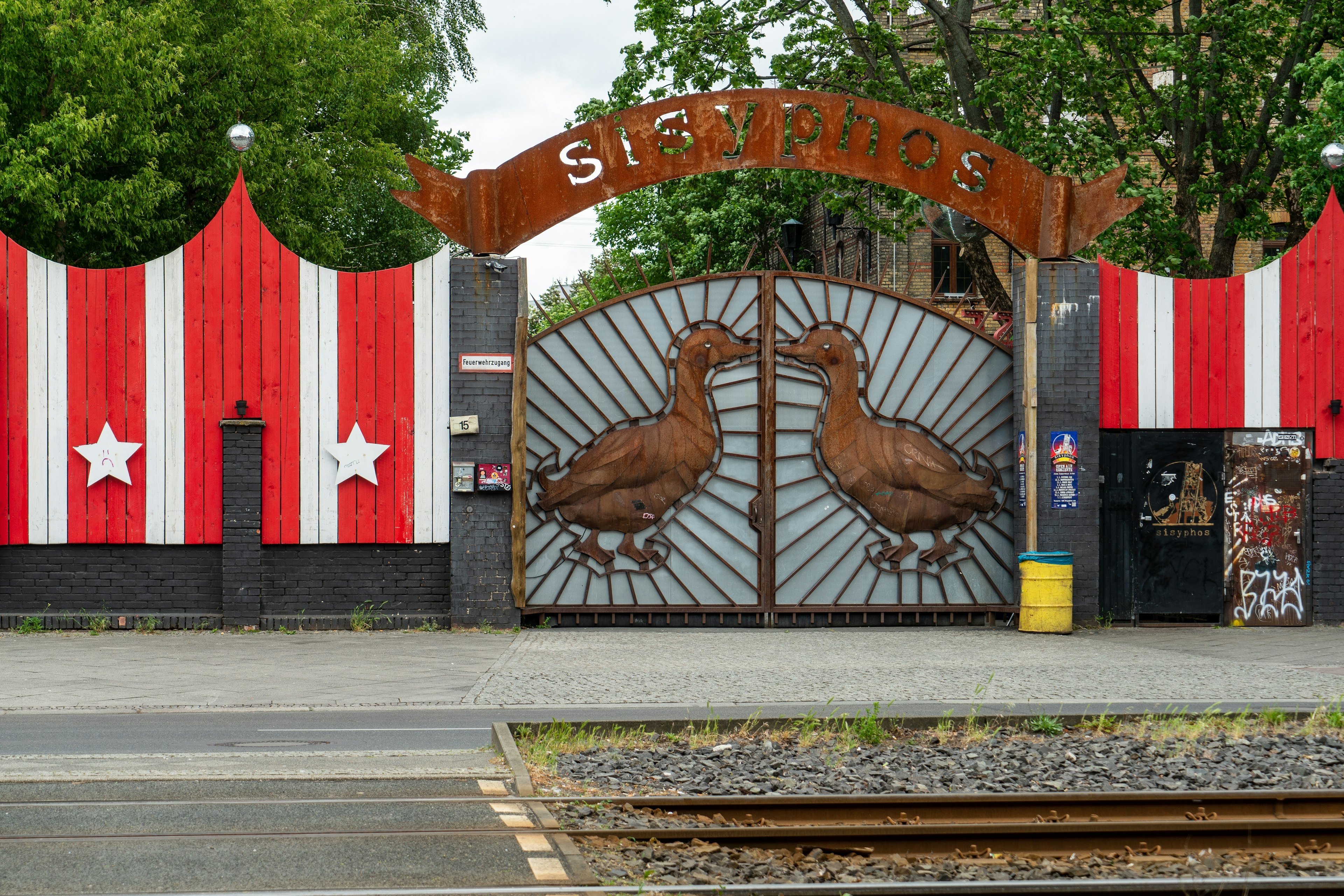 Naktinio klubo išorė su dideliu ženklu virš jo įėjimo vartų, kuriame sakoma "Sisyphos". Vartai yra dekoruoti dviem didelėmis antimis