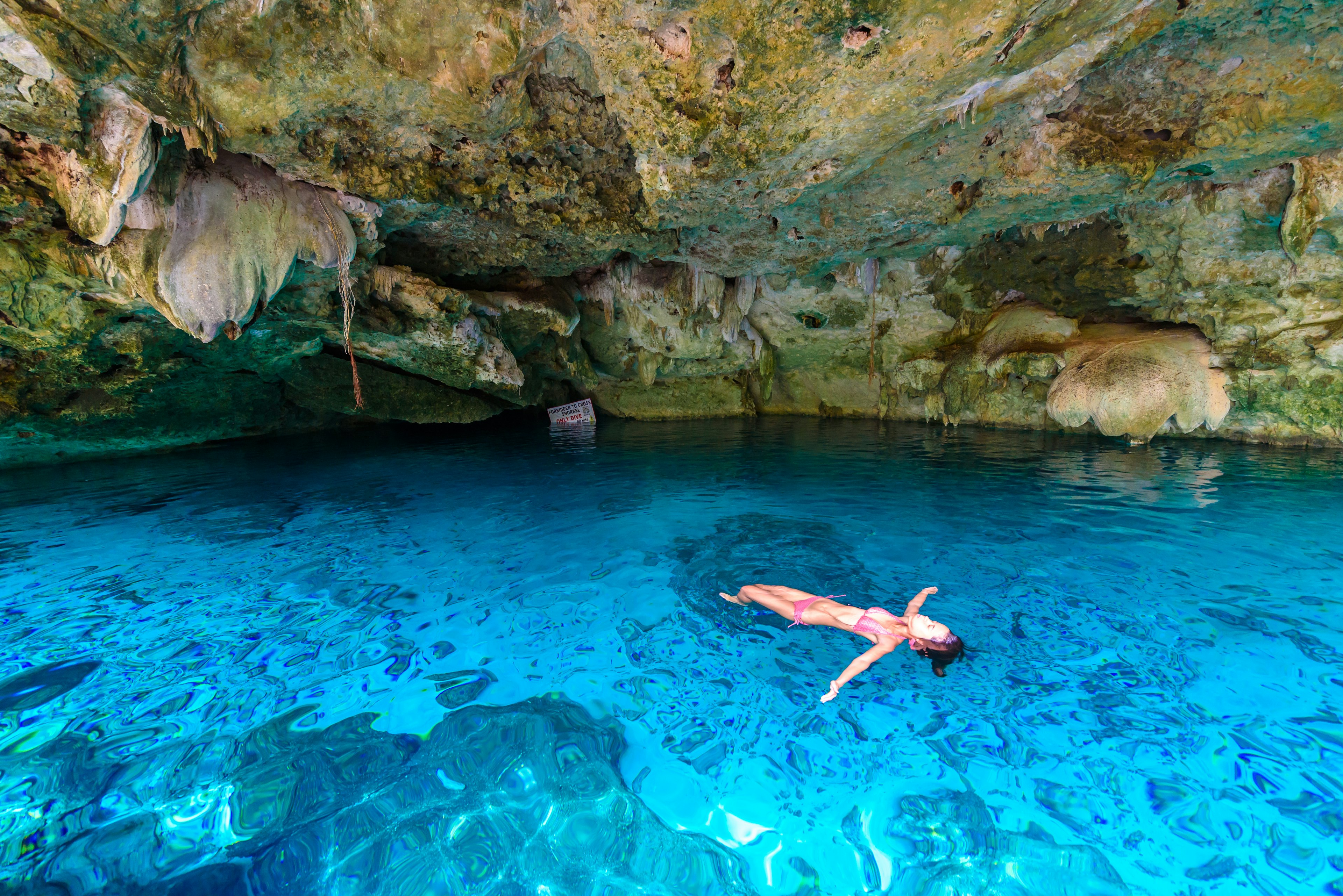 Moteris plūduriuoja mėlynuose vandenyse po cenote urvu.