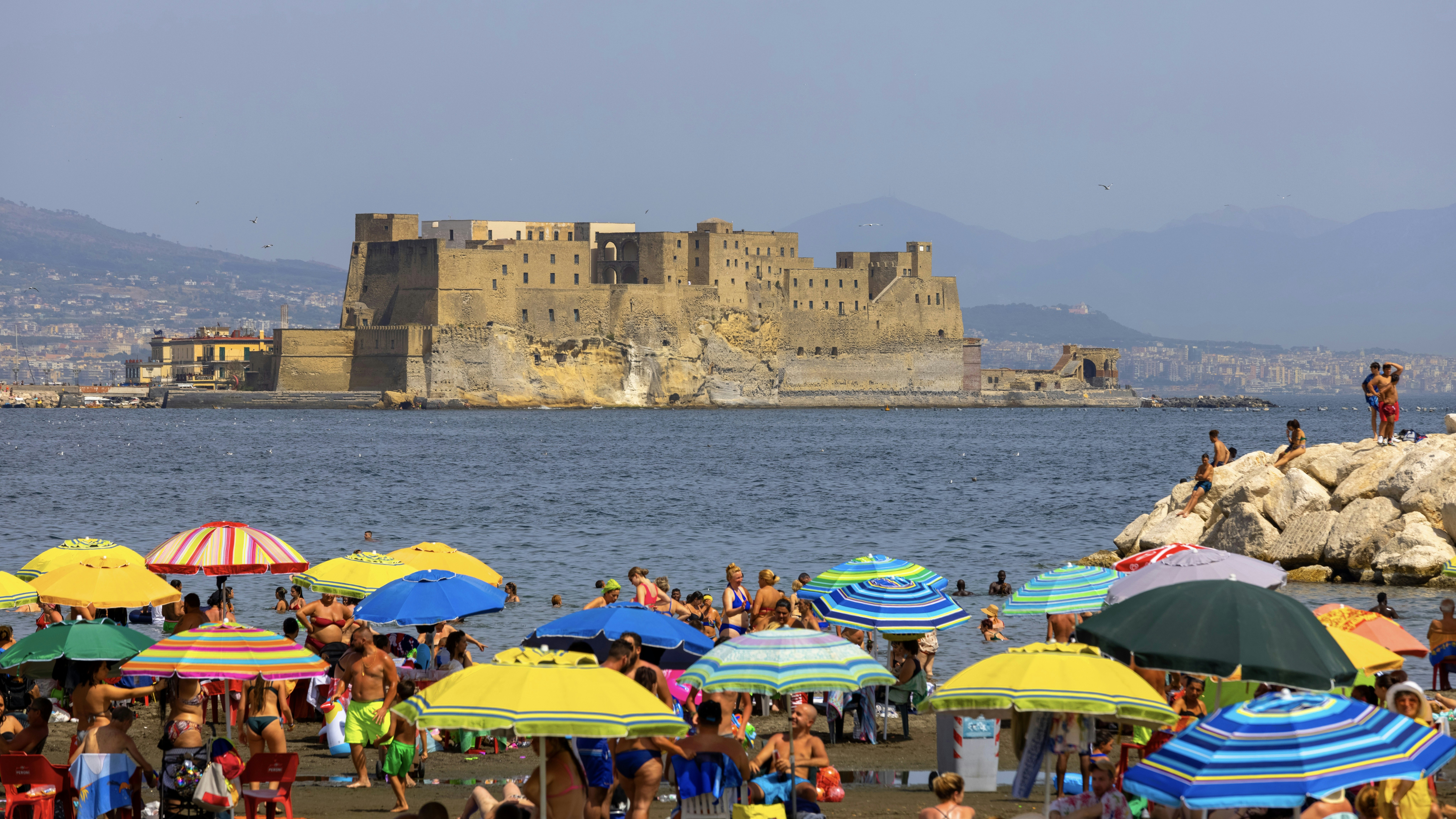 Žmonės, atsipalaidavę Mappatella paplūdimyje prie Tyrhenian jūros šalia bulvaro Chiaa, spalvingi skėčiai, Castel Dell'ovo fone.