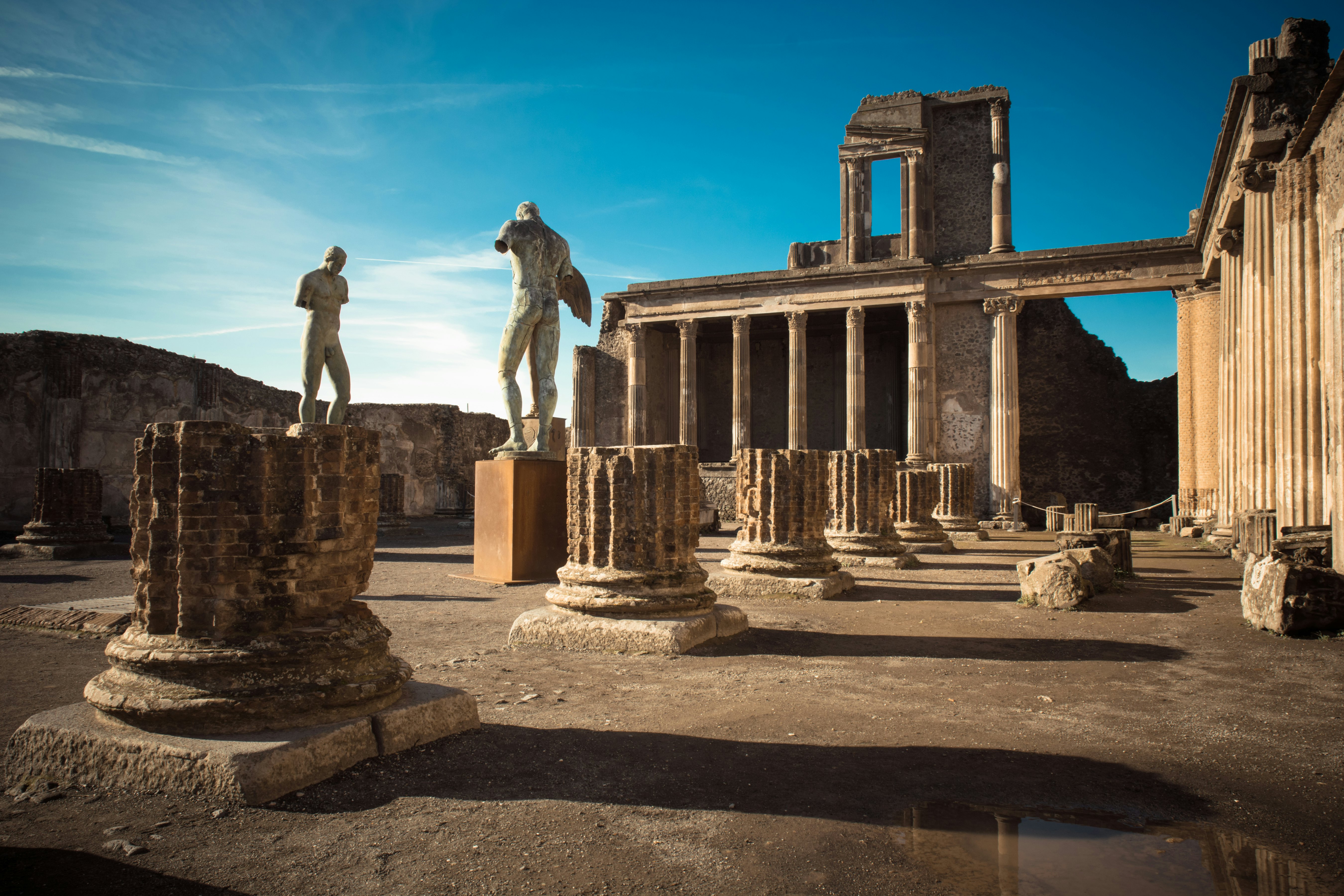 Sugriautos kolonos ir monumentalios statulos kieme, apsuptame kitų kolonų, po mėlynu dangumi su dramatiškais šešėliais