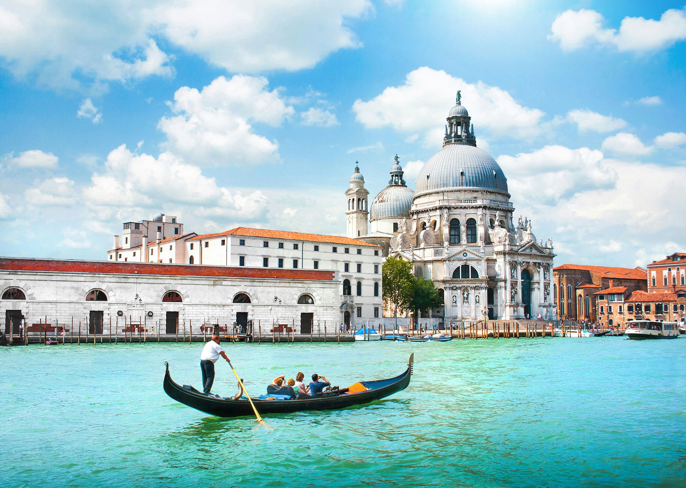 Gondolier eilės juoda gondola su keleiviais ant plačios vandens telkinio. Ant kranto matoma milžiniška, įmantri bažnyčia