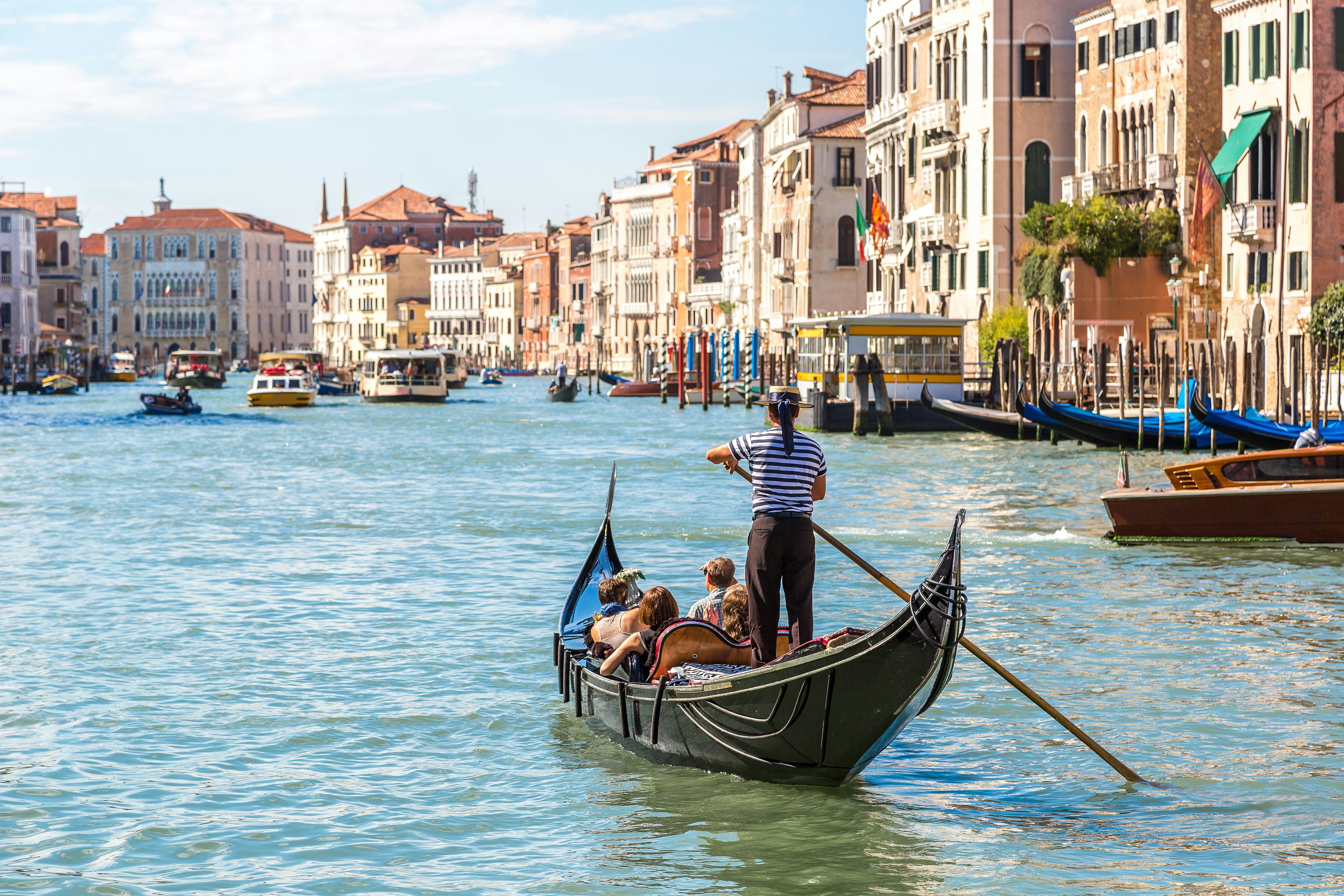 „Gondolier“ saulėtą dieną vairuoja mažą juodą valtį palei kanalą, išklotą istoriniais pastatais