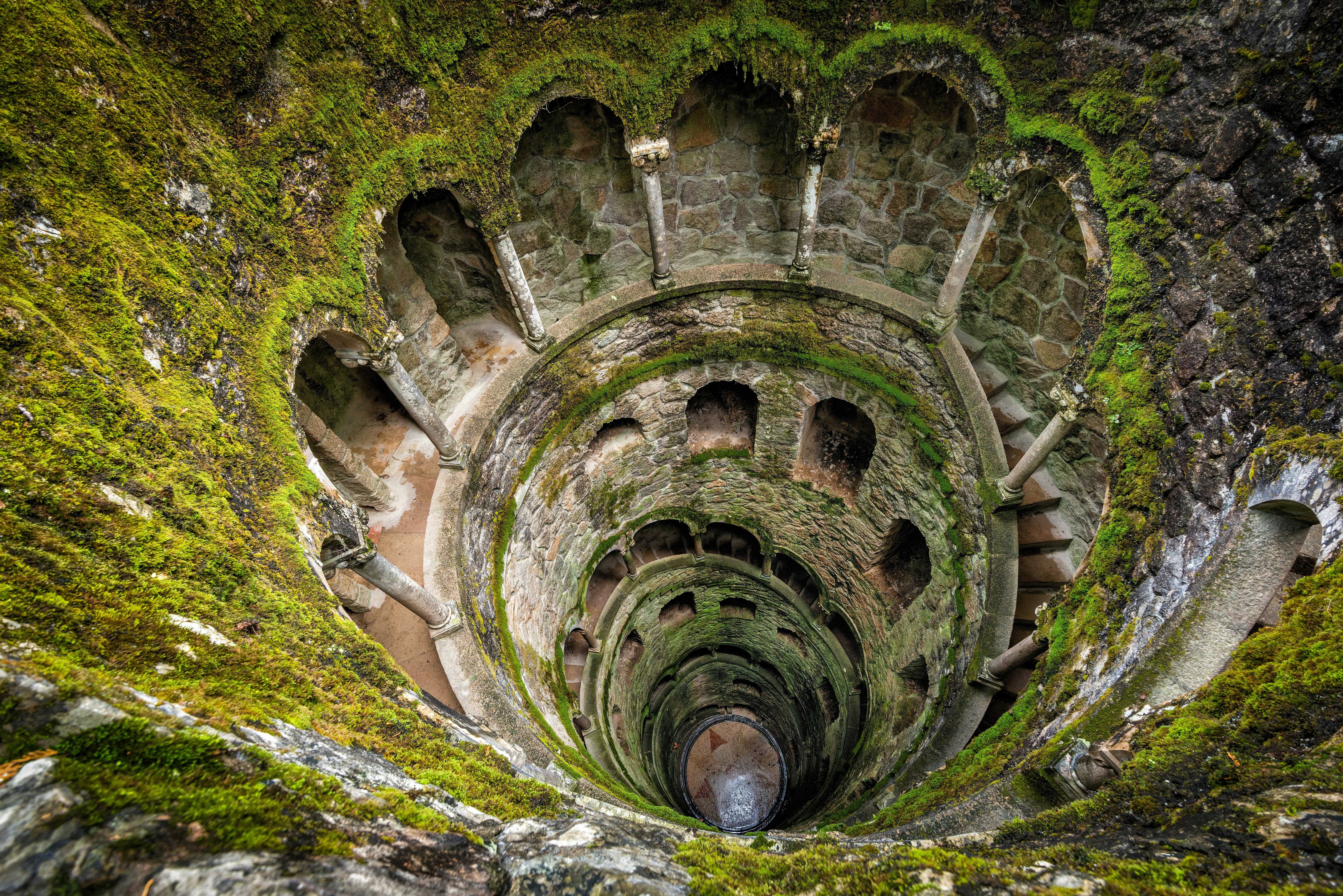 Žvelgiant žemyn į plačią šulinį, su spiraliniais laiptais ir nišomis, kurios sudaro spiralę link dugno. Akmens šonai yra uždengti samanomis.