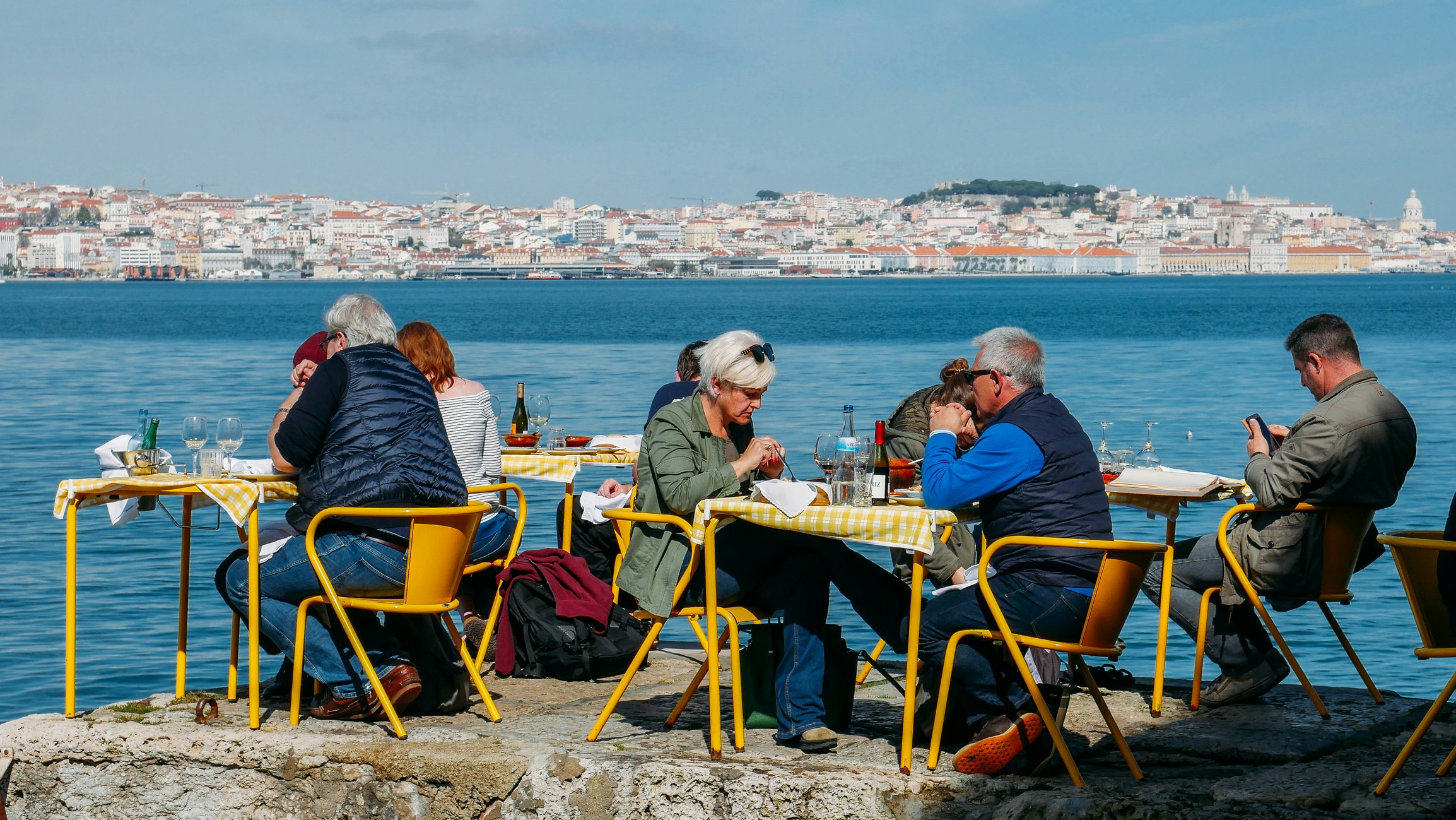 Žmonės, sėdintys prie geltonų restoranų stalų, esančių Tagus upės krante, Kakiluose, o fone - centrinės Lisabonos miesto vaizdas