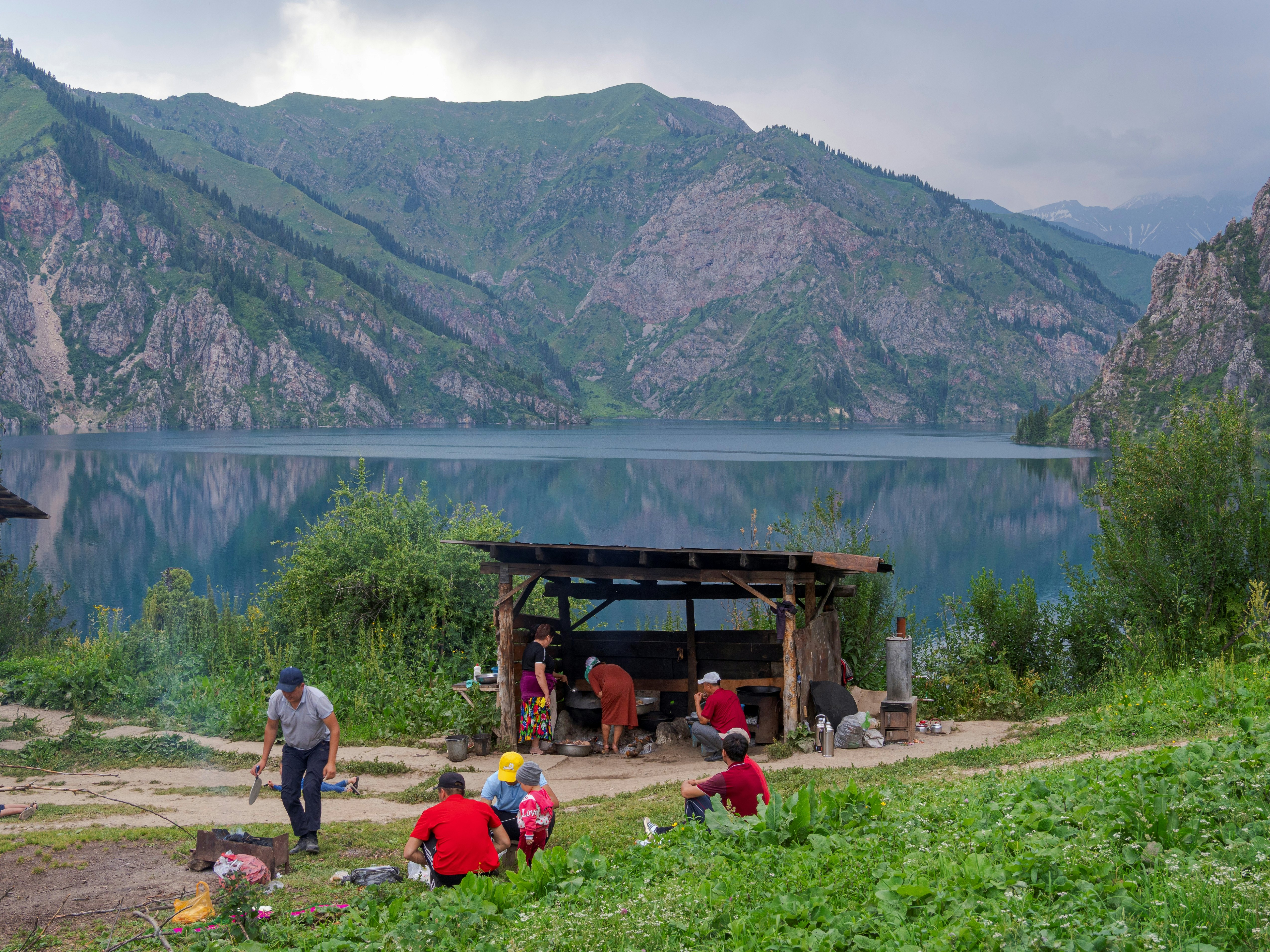 Vietiniai turistai paruošia kepsninę prieglaudoje prie ežero su medžių dengtais šlaitais, kylančiais iš visų pusių, Sary-Chelek ežero, Kirgizstano ežeras