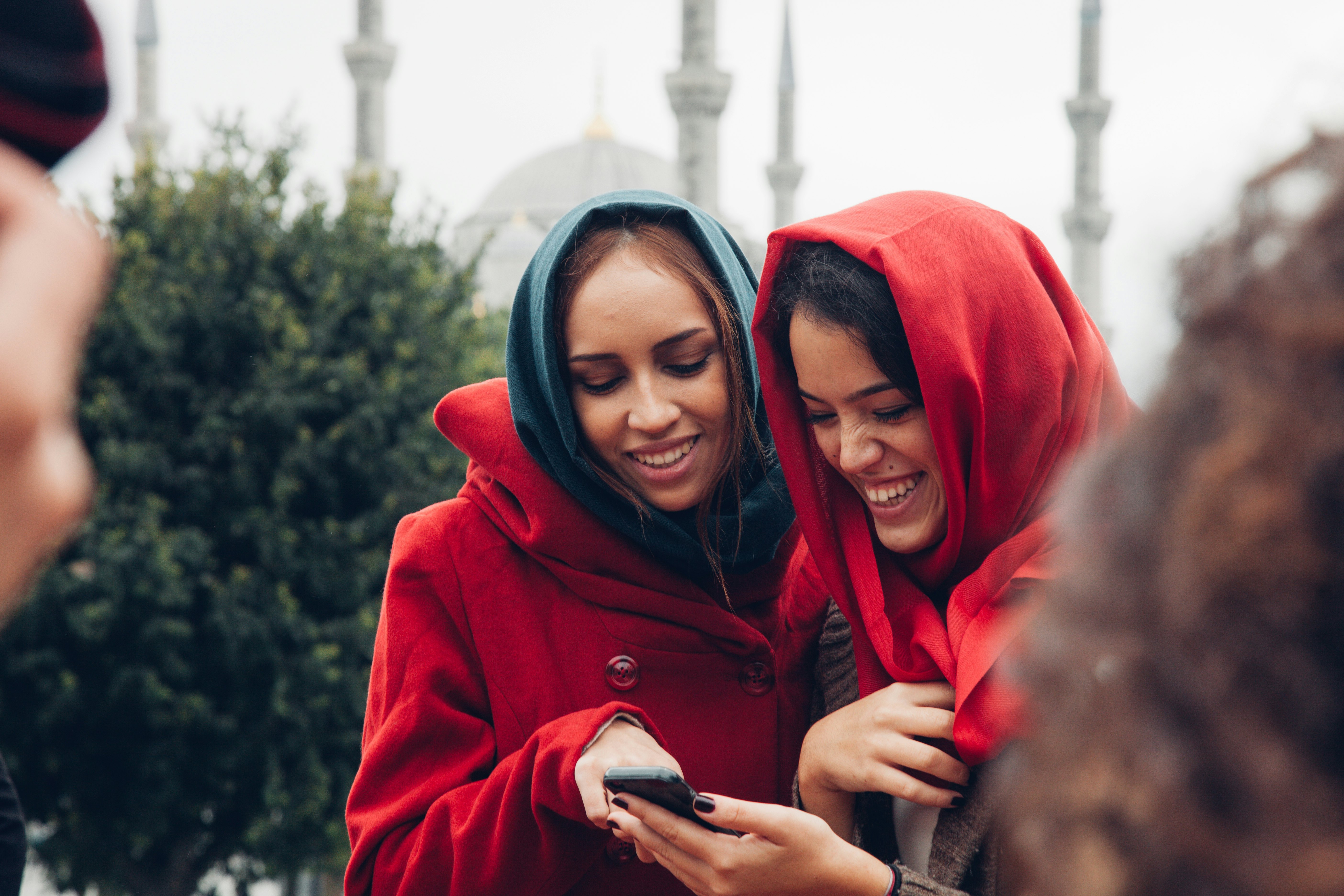 Dvi jaunos moterys galvos viršeliuose juokiasi, kai žiūri į telefoną šalia Mėlynosios mečetės Stambule, Türkiye