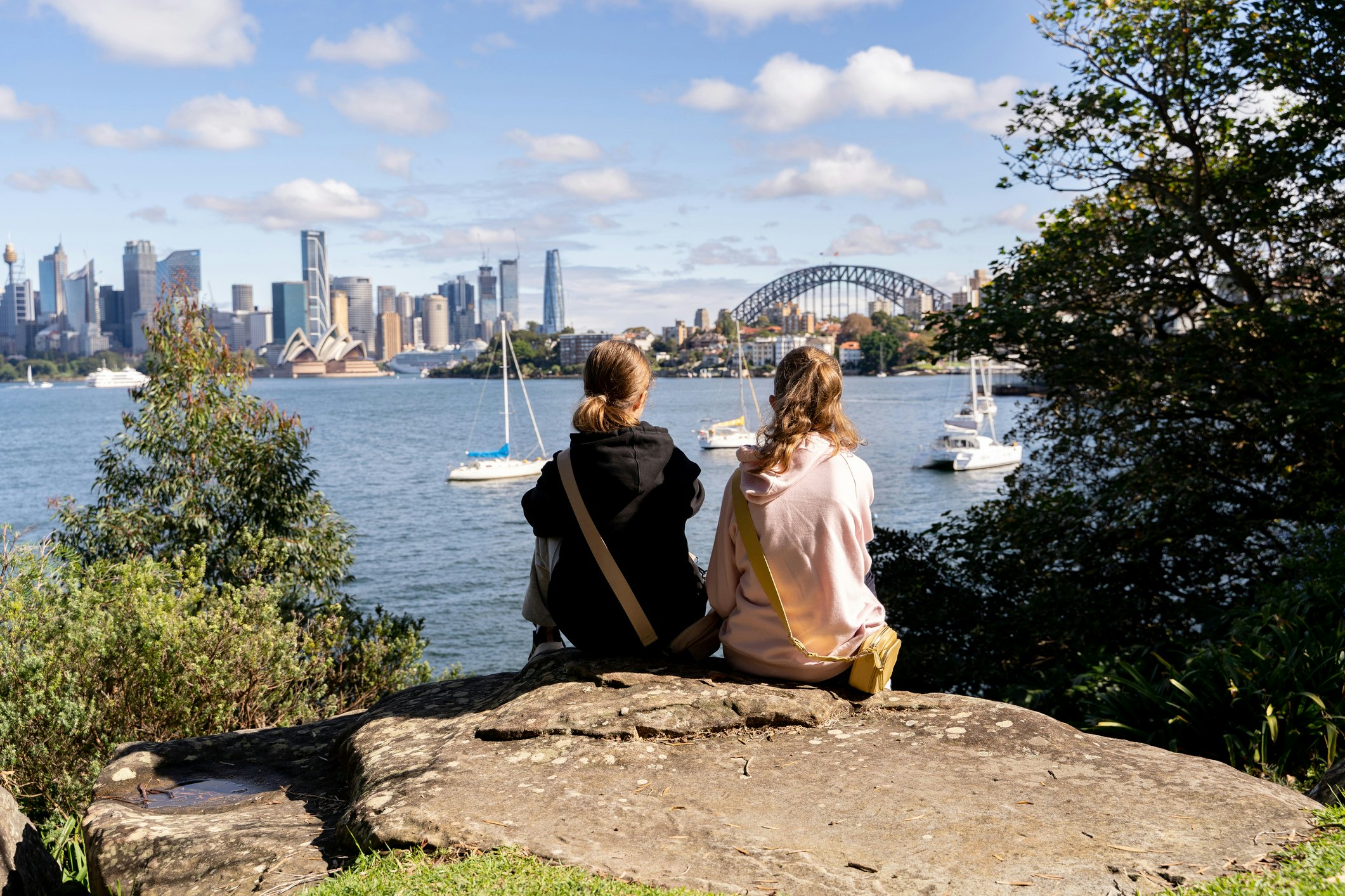 Two girls sit on a rock and look out over an iconic harbor, with a white opera house and a large bridge