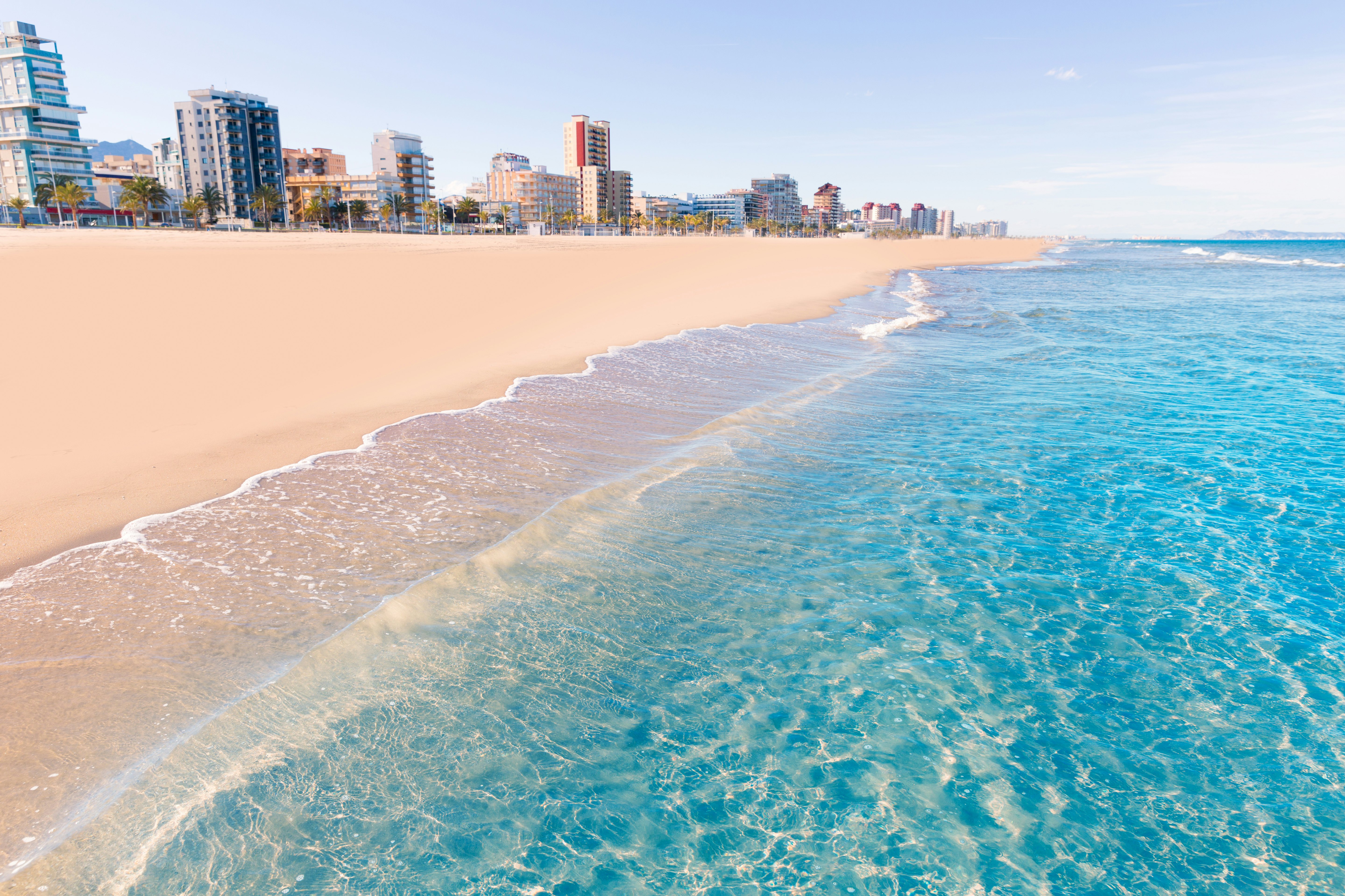 Aiškus, mėlynas Viduržemio jūros plovimas įstrižai supjaustomas prieš minkštą, rausvą „Playa de Gandia“ smėlį Valensijoje, Ispanijoje su keliais bokštų blokais, kylančiais fone