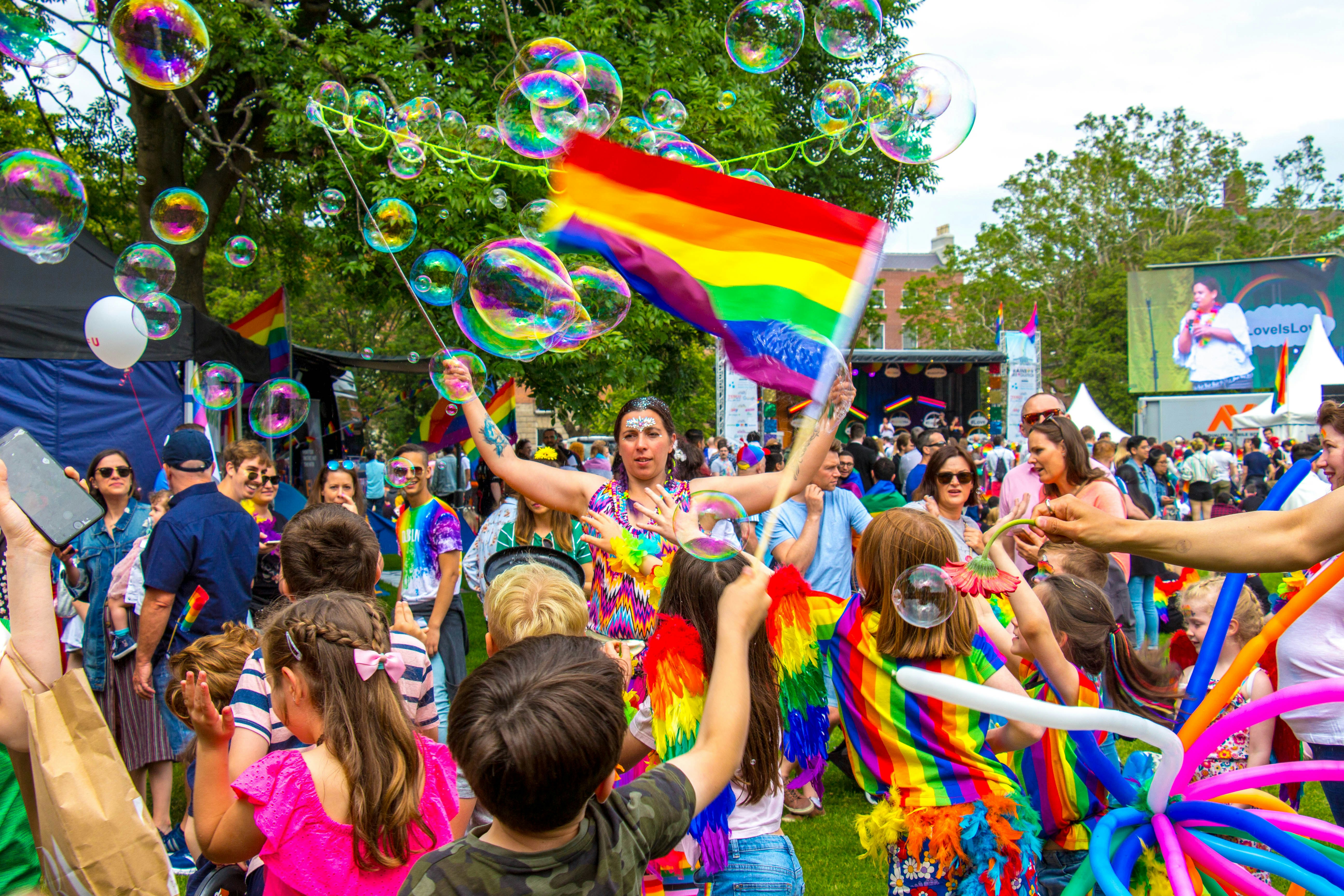 Grupė žmonių, suaugusių ir vaikų, pasipuošusių ryškiais drabužiais švenčia Pride festivalį Dubline birželio mėnesį, mojuoja vėliavomis ir pučia burbulus
