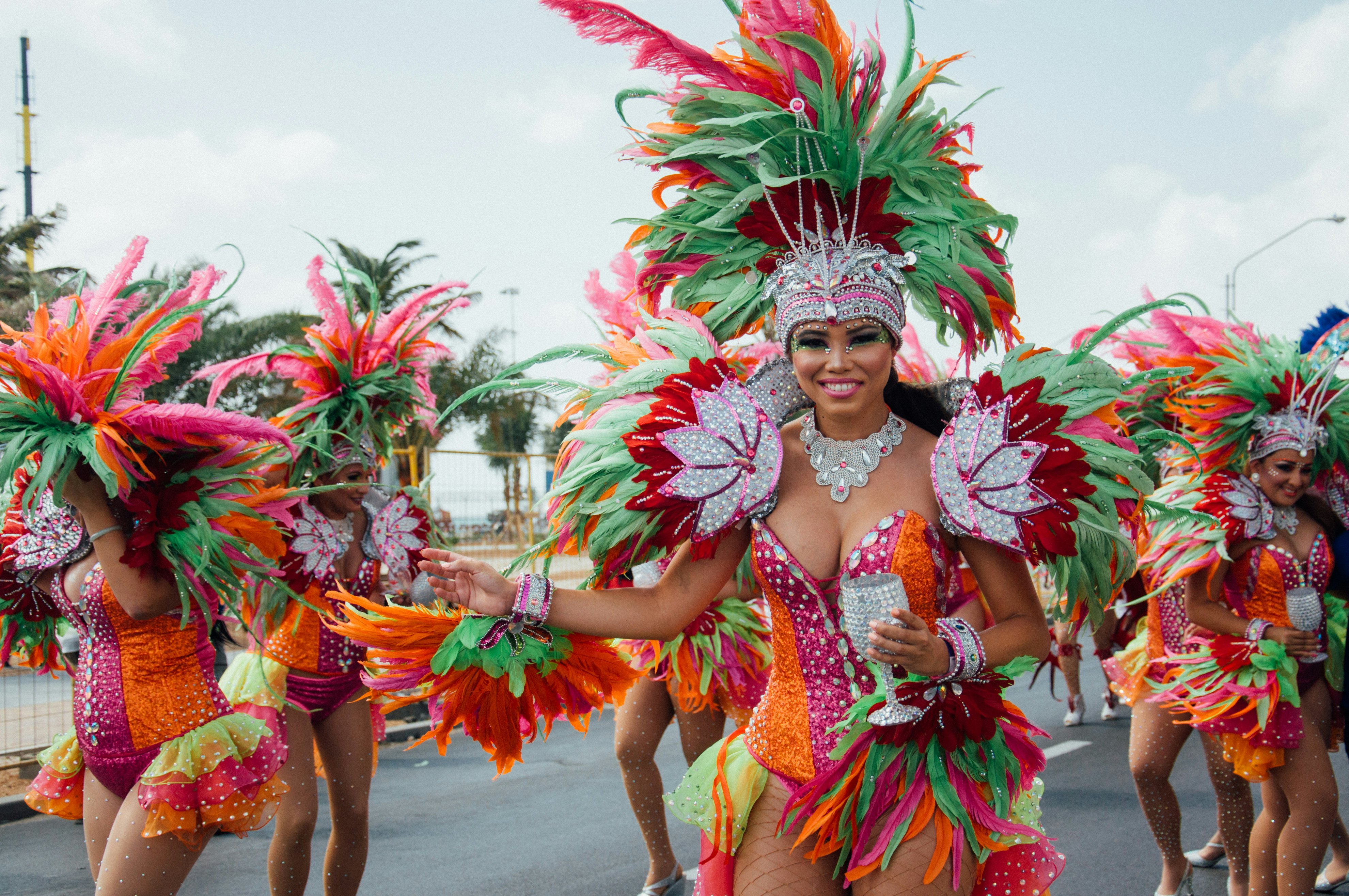 Moteris su ryškiai oranžine žalia trumpa suknele ir didelė dekoratyvinė karūna su plunksnomis, einančiomis ir šypsodamasis karnavalo procesijos metu, Aruba