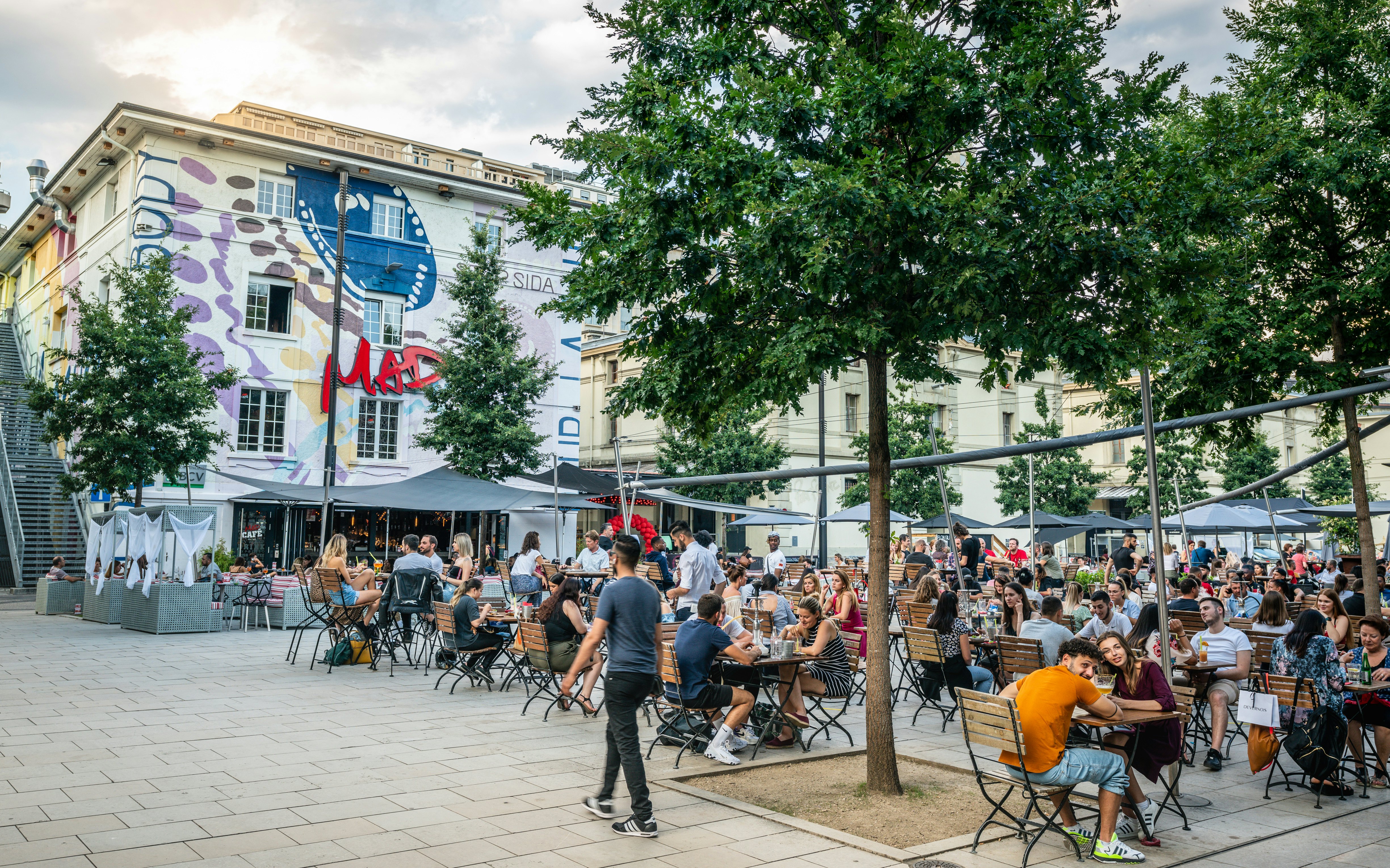Vyras vaikšto prie jaunų žmonių, sėdinčių prie stalų lauko terasoje ant miesto aikštės vasarą