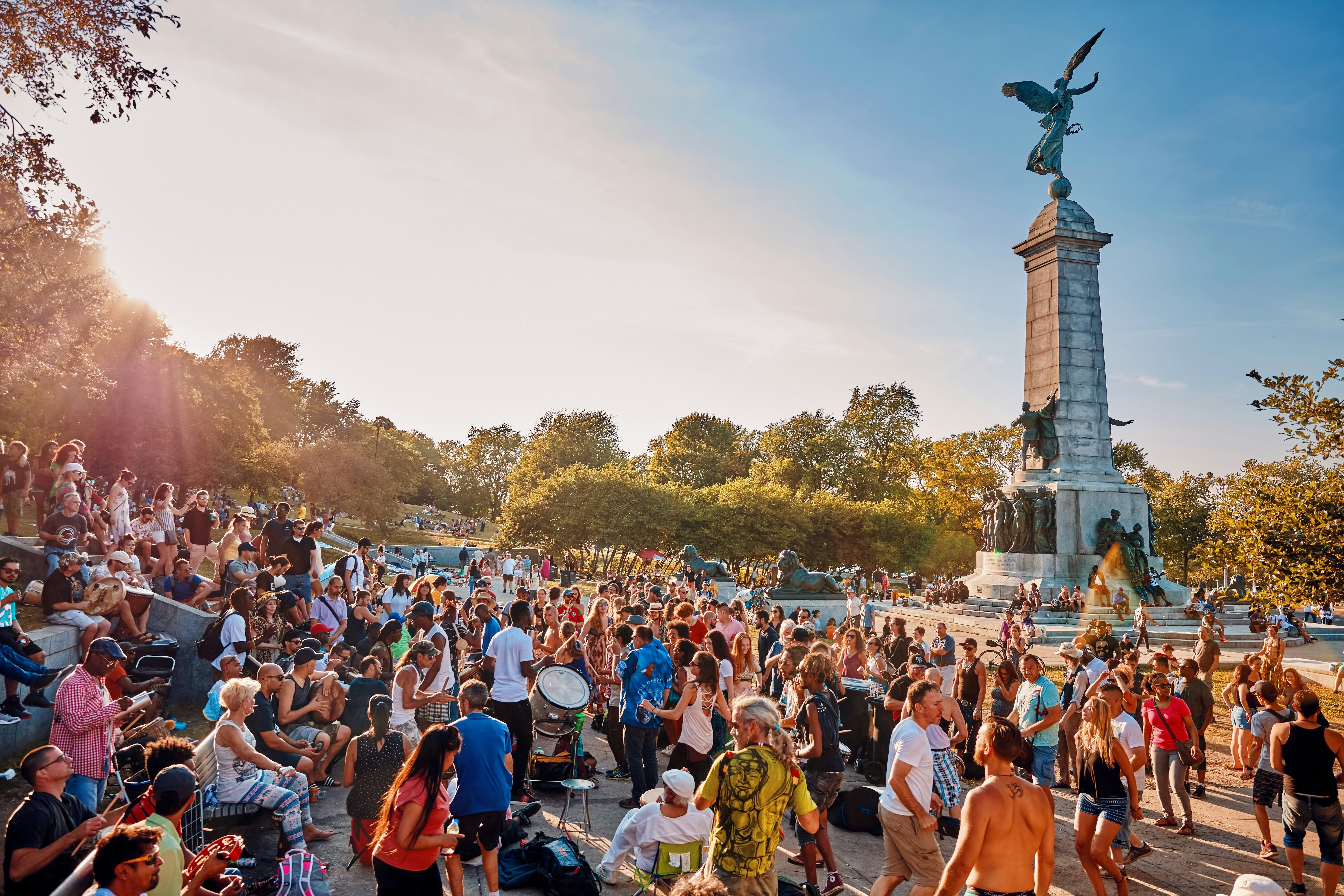 A diverse crowd of people gathered and cheering around a tall monument in a park. Some people are beating drums