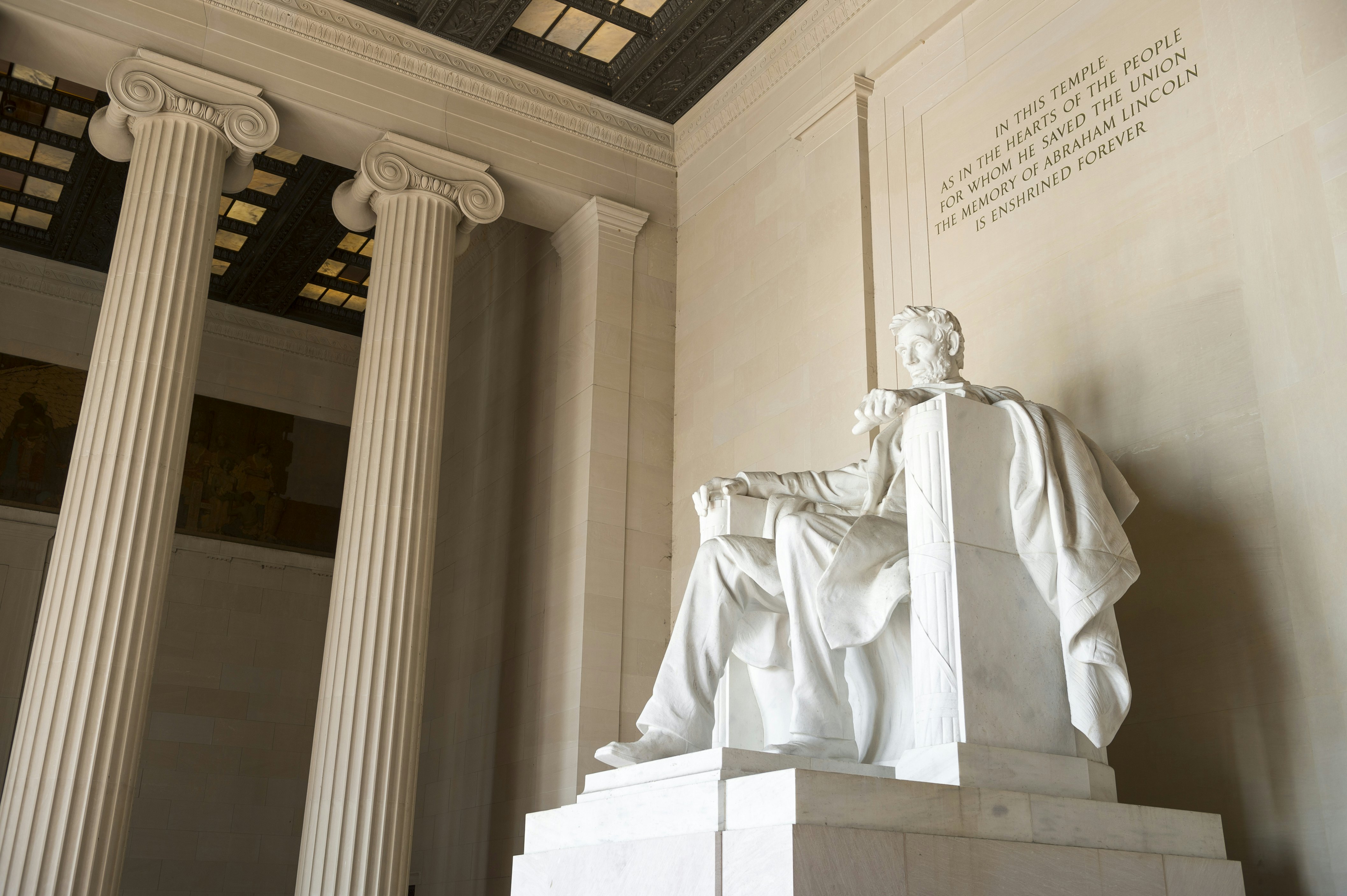 A vast white-stone sculpture of a man sat on a chair in a space with decorative columns. Text engraved into the wall above says