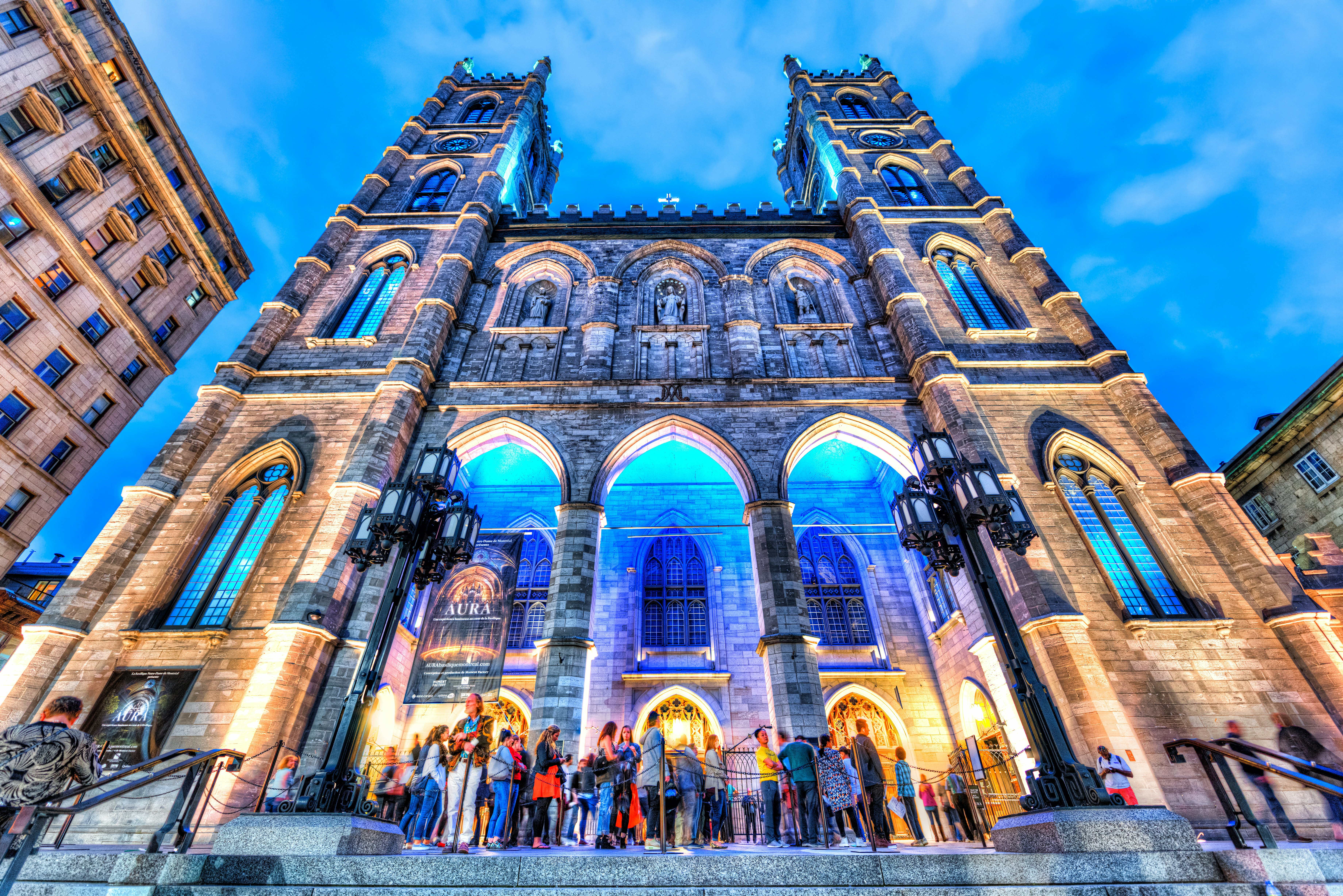 People gather outside a Gothic cathedral in the evening, which is lit up with pale blue lights