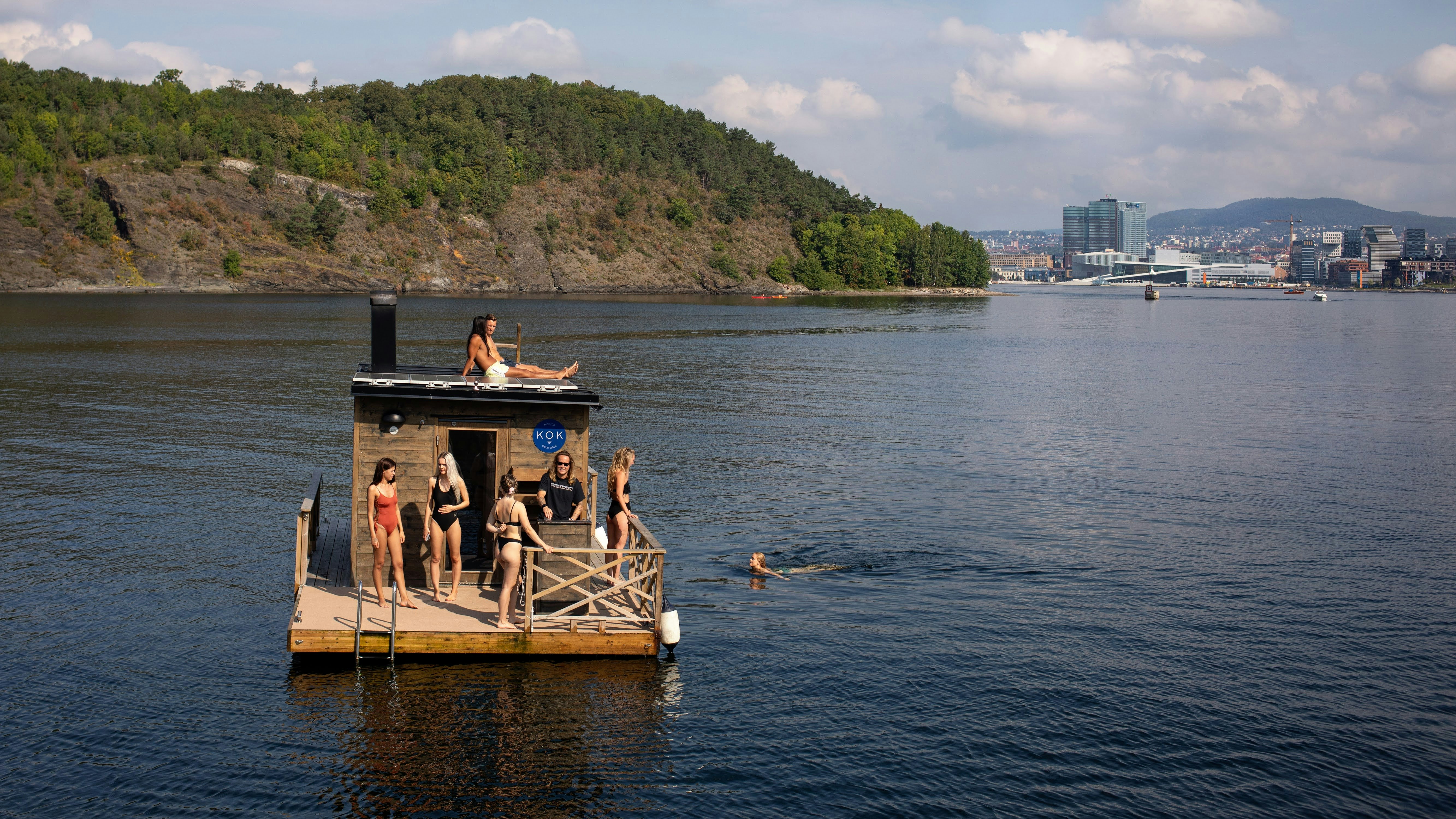 Žmonės mėgaujasi plaukiojančia KOK sauna fjorde šalia Oslo. Vienas plaukia, kiti deginasi ant stogo ir denyje. Vairuotojas vilki juodą marškinėlius.