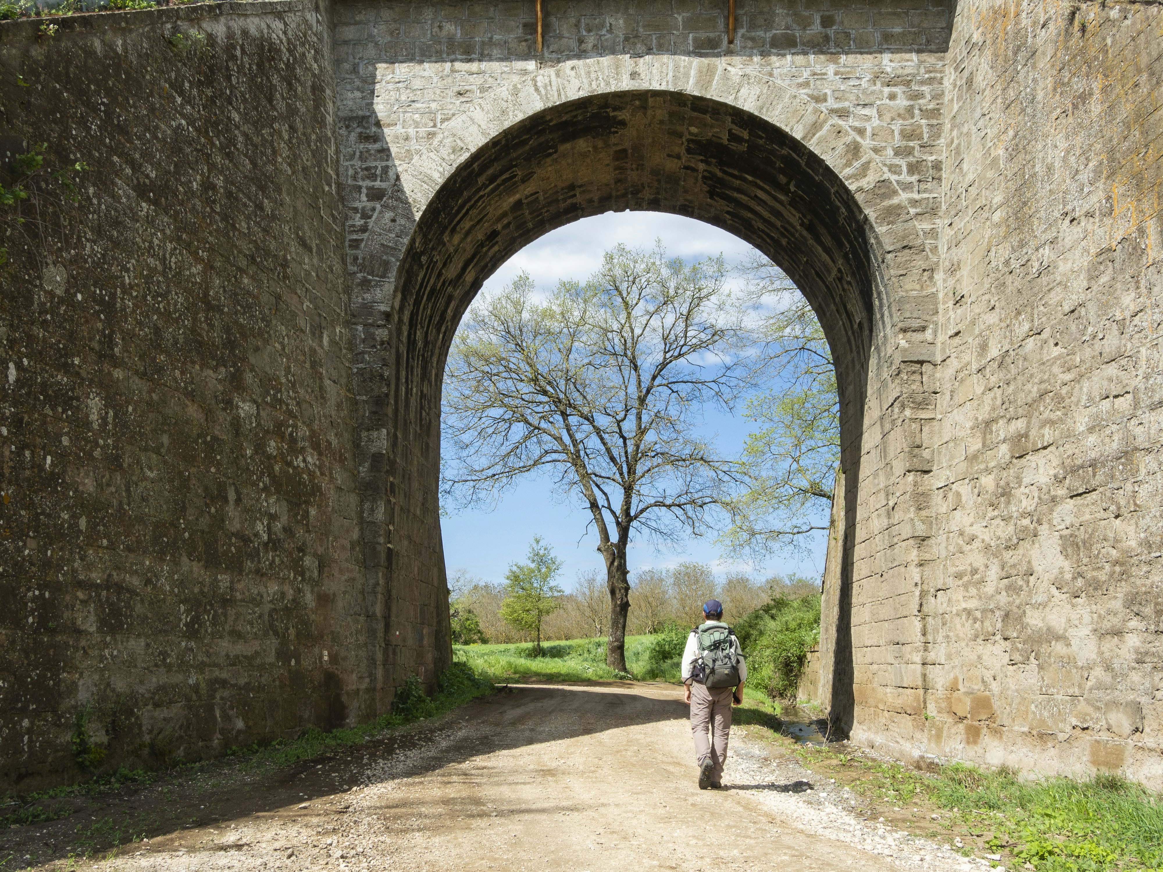 Piligrimas su kuprine eina pro akmeninį arką šalia Vetralla, Via Francigena trasoje