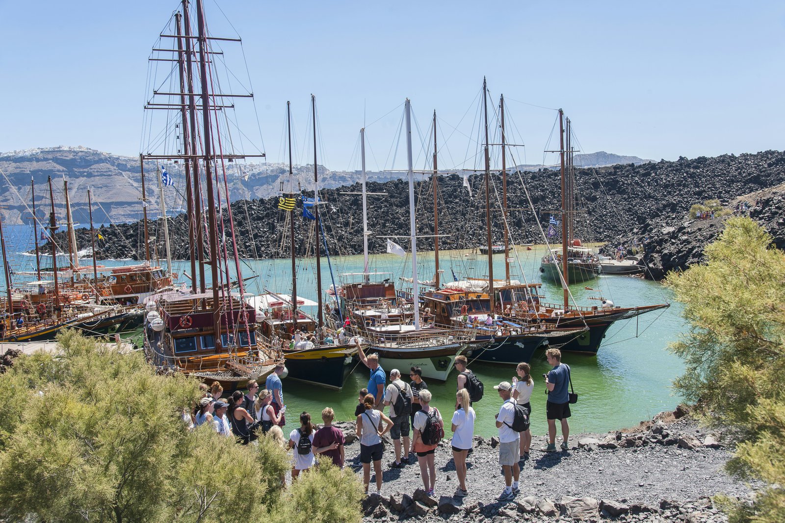 A view of people standing near the Nea Kameni volcano with boats and trees.