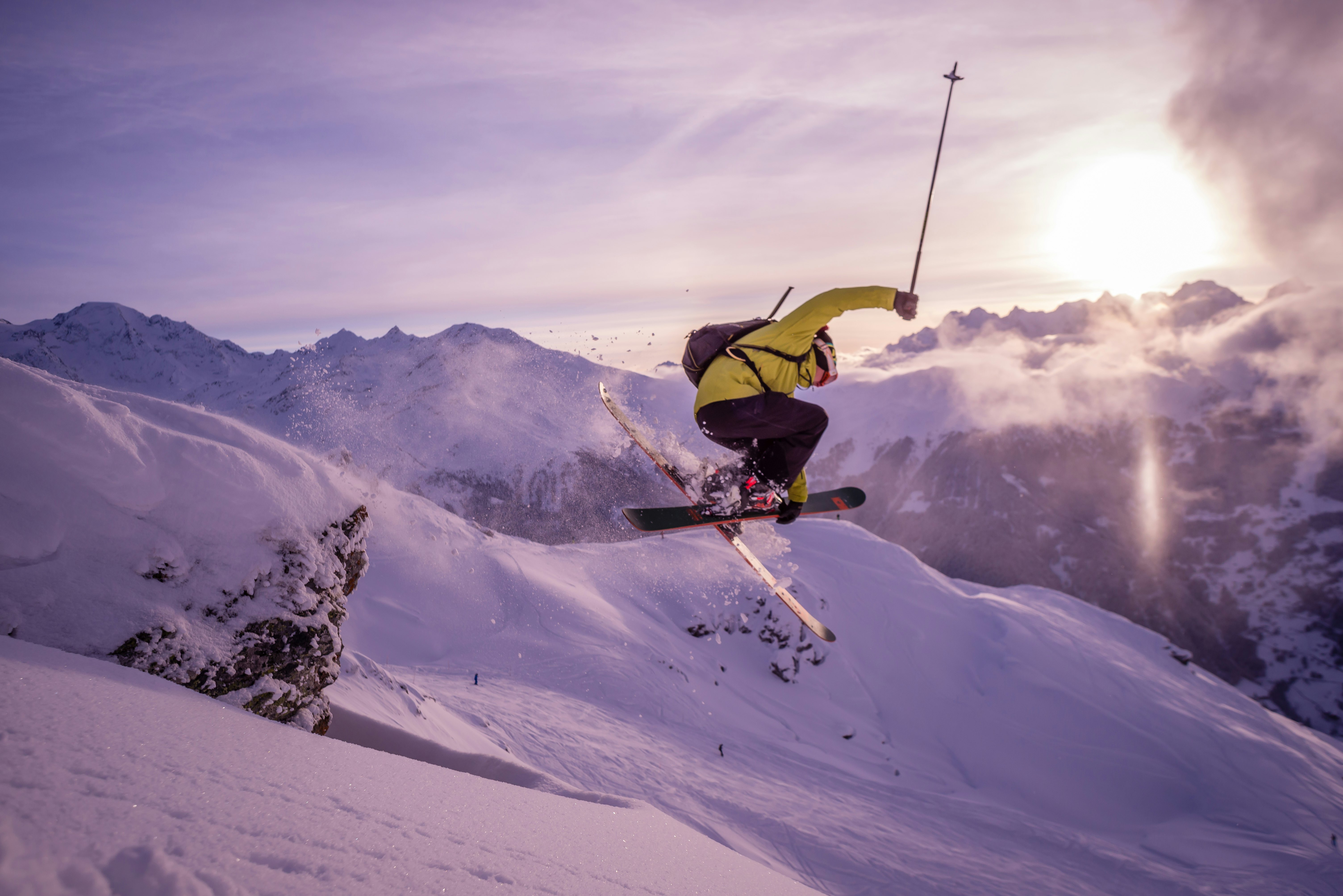 Slidininkas šoka ore virš Verbier kalnų. Viršūnių fone matosi sniegas ir leidžiantis saulė.