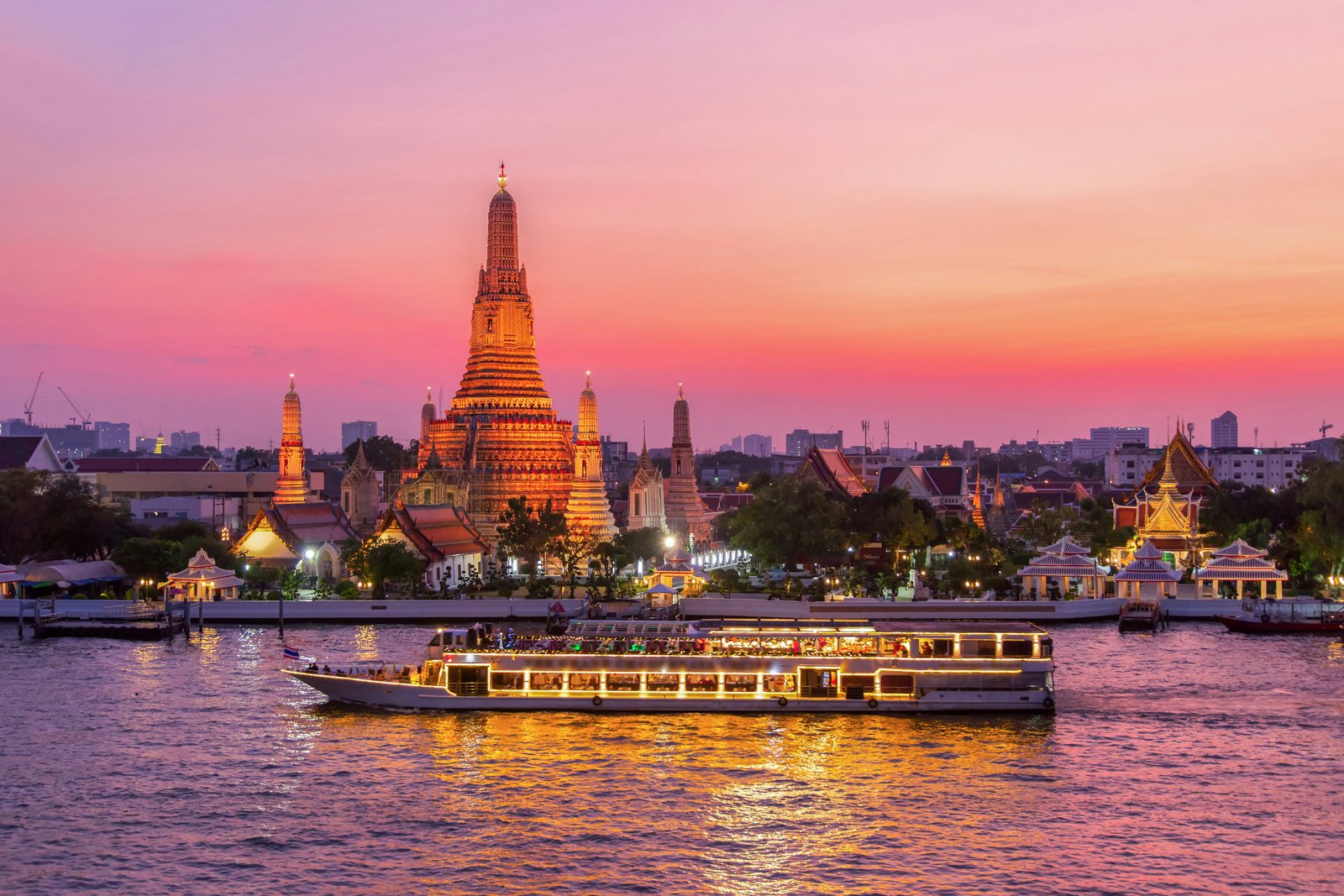 A boat with lights on its windows passes a tall stupa by a river. The scene is bathed with pink and orange sunset light.