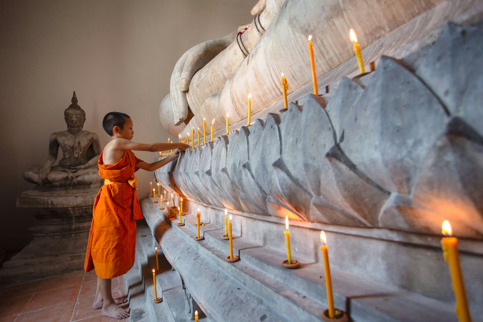 A young monk in a saffron robe lights a candle at the base of a Buddha statue in a temple.