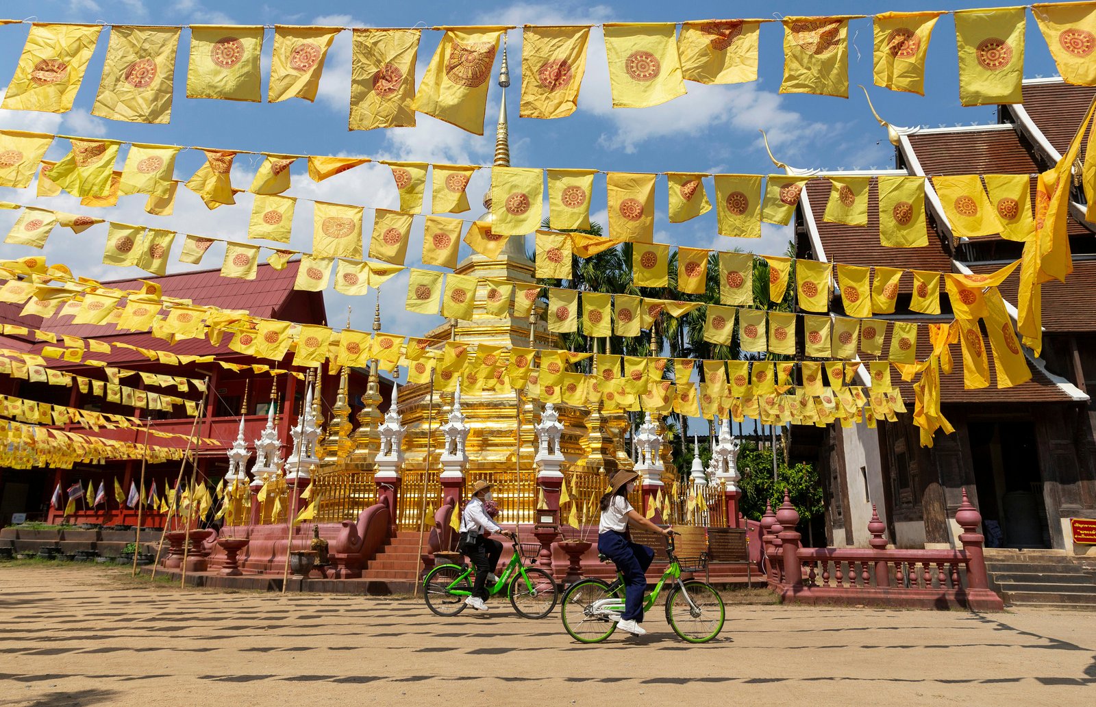 Two people on bicycles pedal past the gold stupa of a temple. Numerous yellow flags hanging from strings flutter overhead.