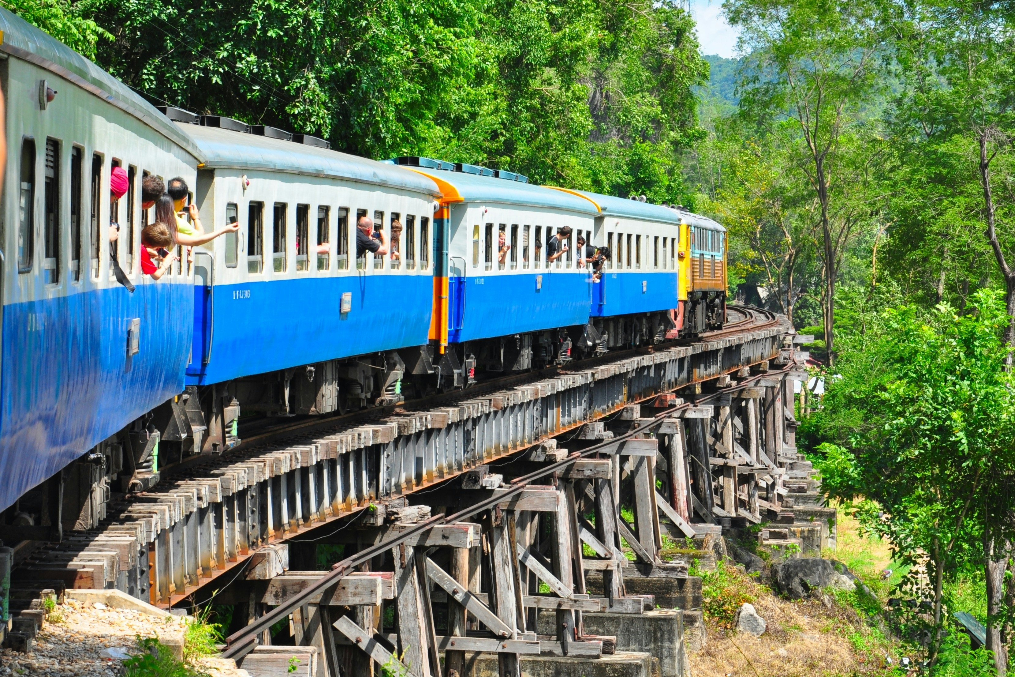 Turistai išlindę pro traukinio langą žvalgosi keliaudami tiltu per Tailando kraštovaizdį