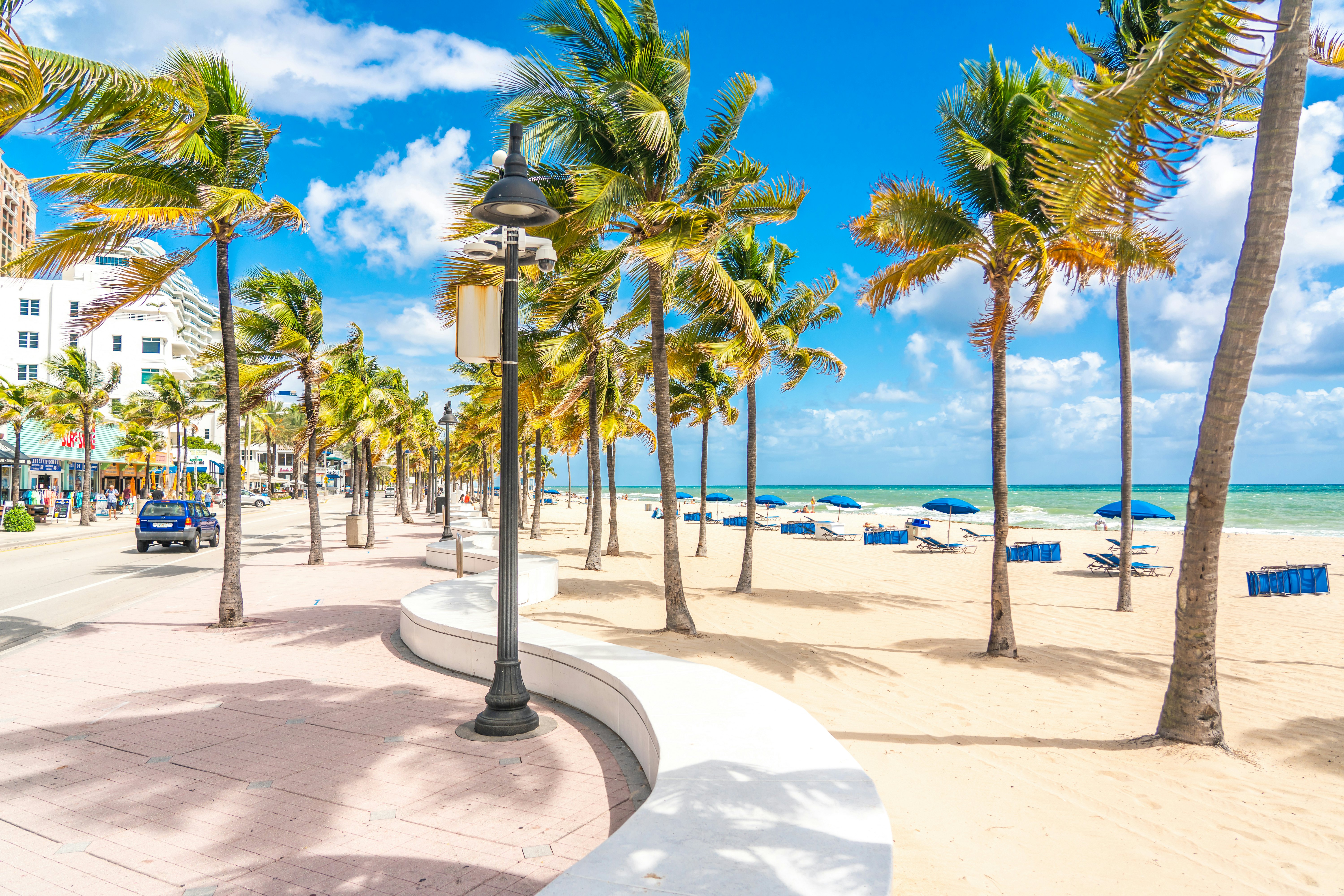 The seafront beach promenade in Fort Lauderdale is lined with palm trees, while blue sun loungers are laid out on the white-sand beach beyond.