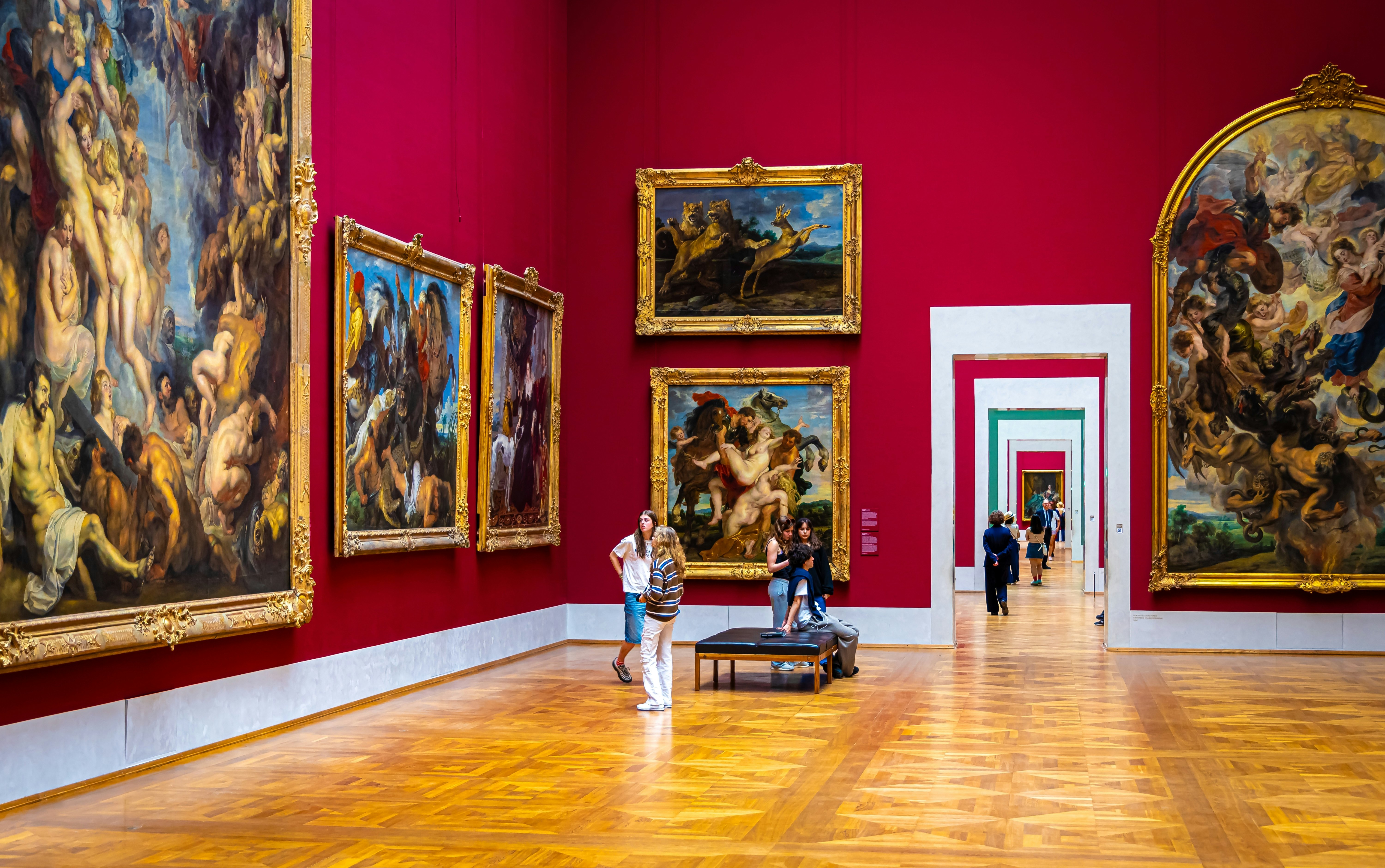 Lankytojai vaikšto galerijomis su raudonomis sienomis, puoštomis didžiuliais paveikslais Alte Pinakothek muziejuje, Miunchene