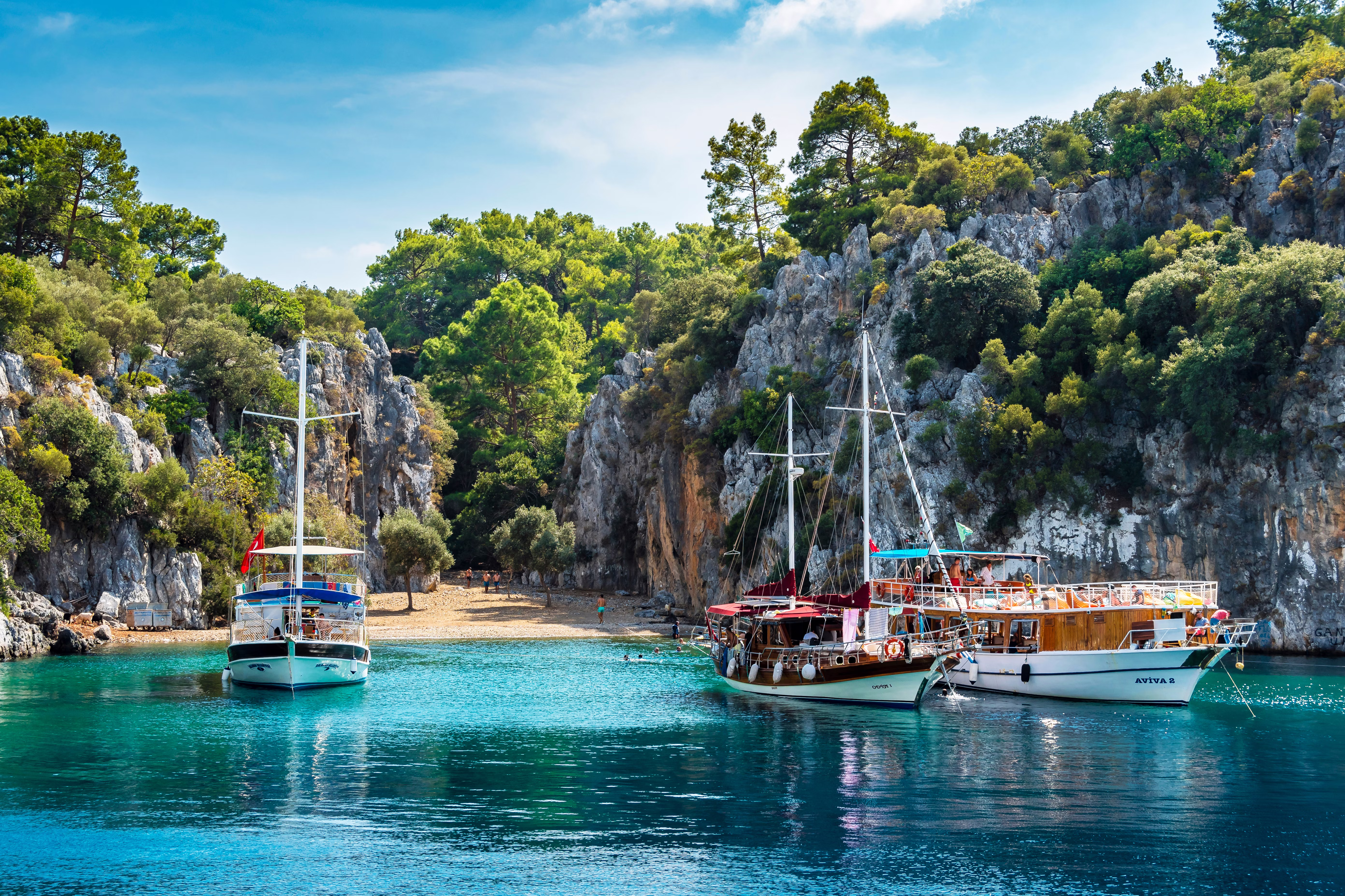 Ships docked in a small cove in Türkiye with turquoise water and a golden beach; there are large rocky cliffs on either side of the beach, and green trees beyond.