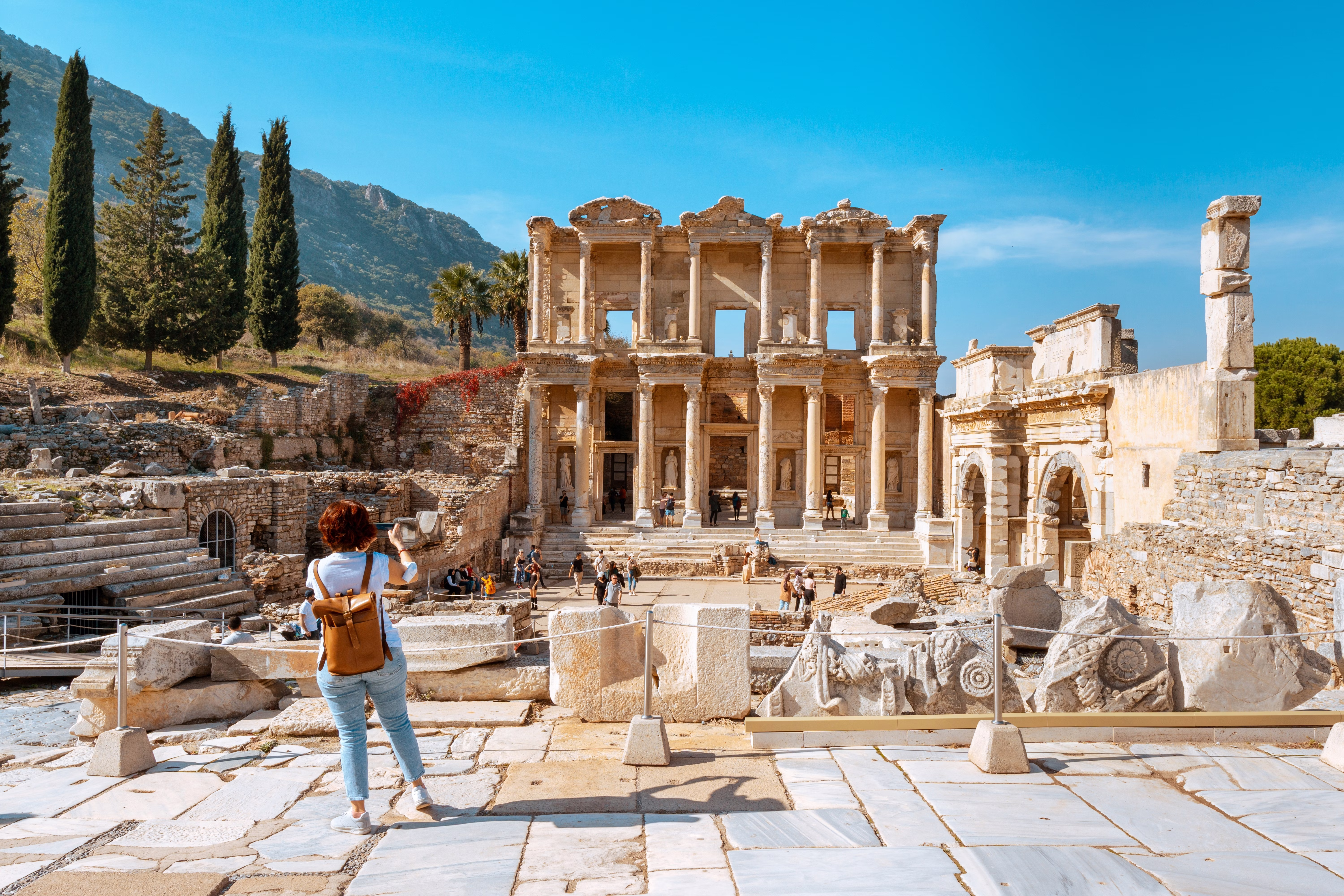 A visitor wearing jeans and a leather backpack takes photos of ancient ruins in Türkiye.