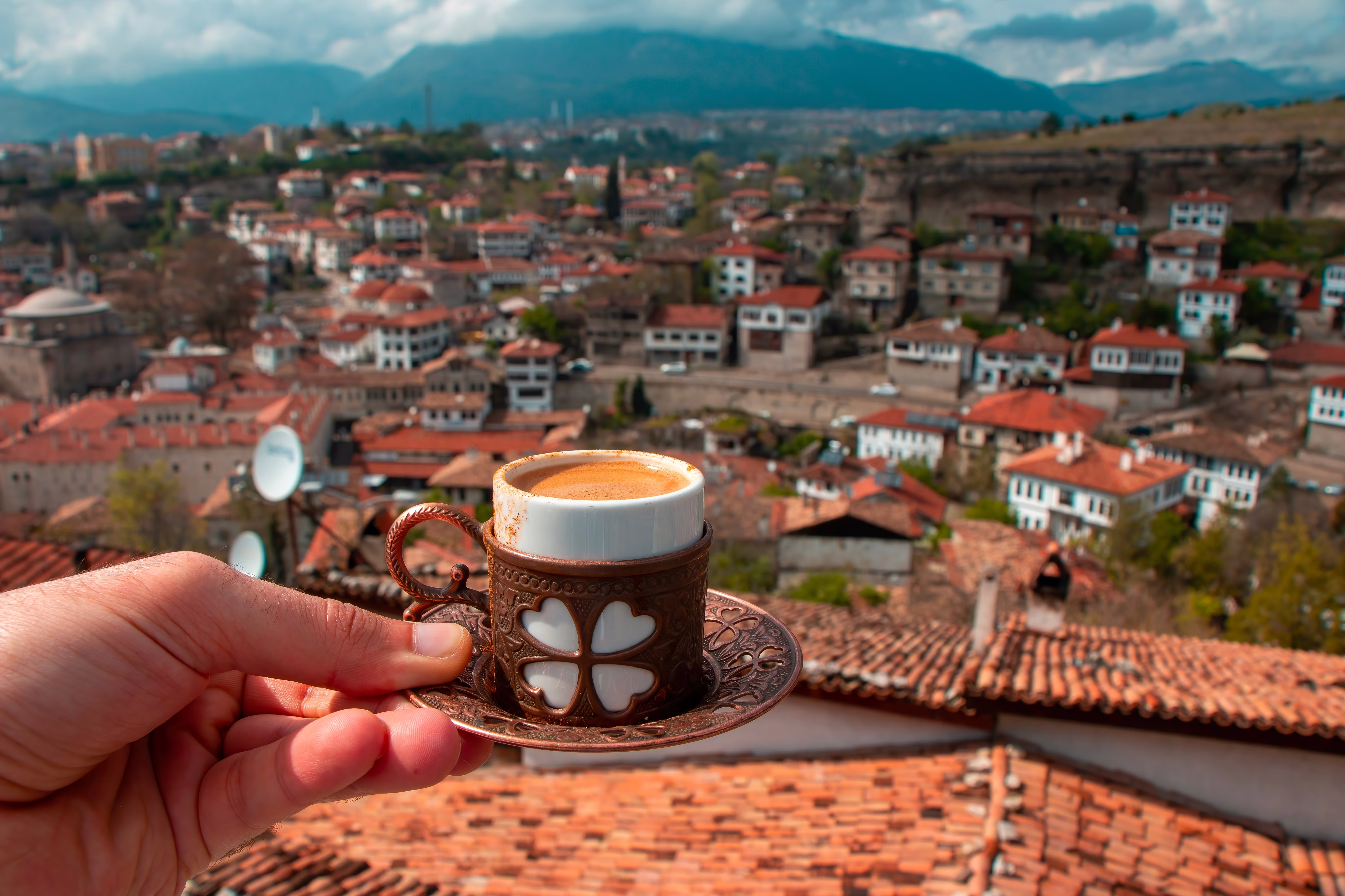 A hand holds out a small cup of coffee on a saucer with a view of a city with low-rise houses with red roofs