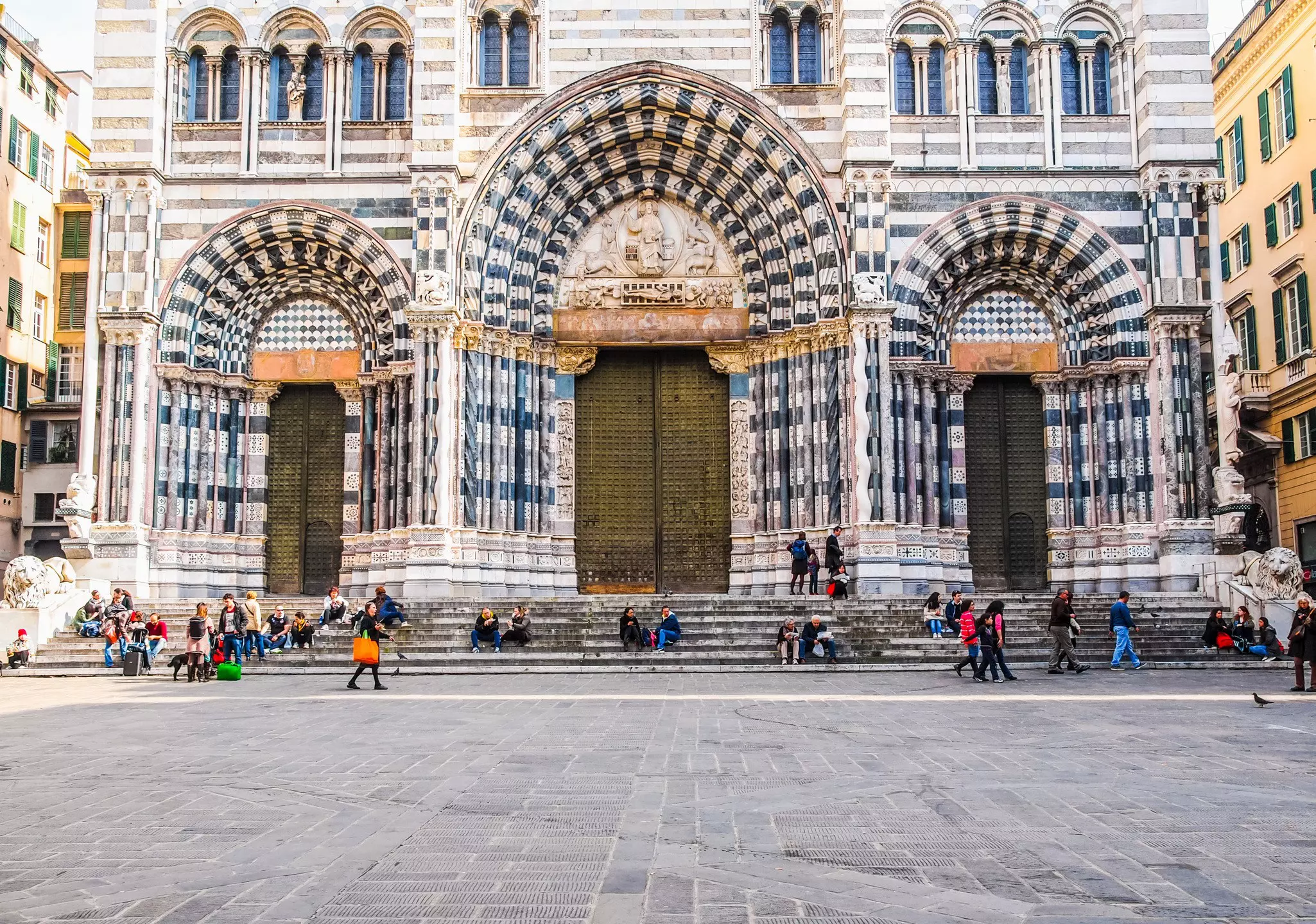 People sit on the steps at the front of a Gothic and Romanesque-style cathedral.