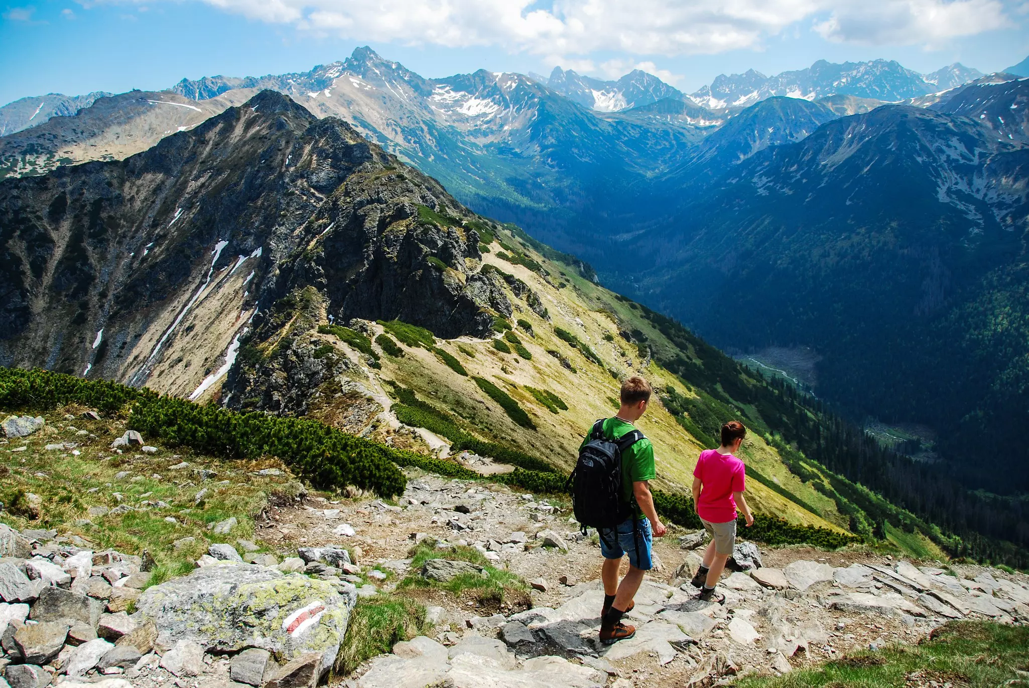 Two hikers walk down a steep rocky path into a mountain valley in the summer; mountains in the distance have small patches of snow.