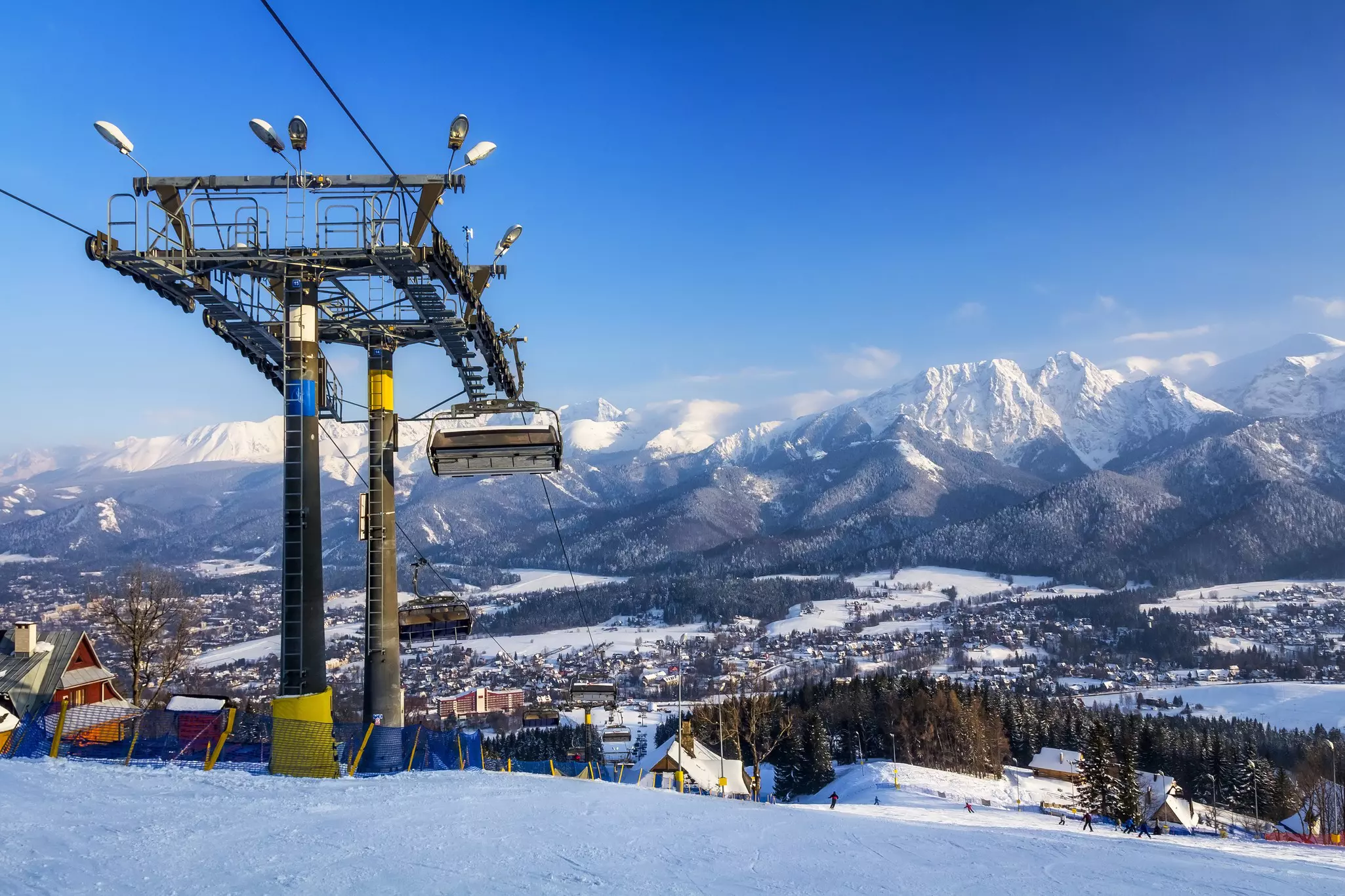 A ski lift at the top of a hill; a snow-covered town is visible in the valley and a mountain range is on its other side.
